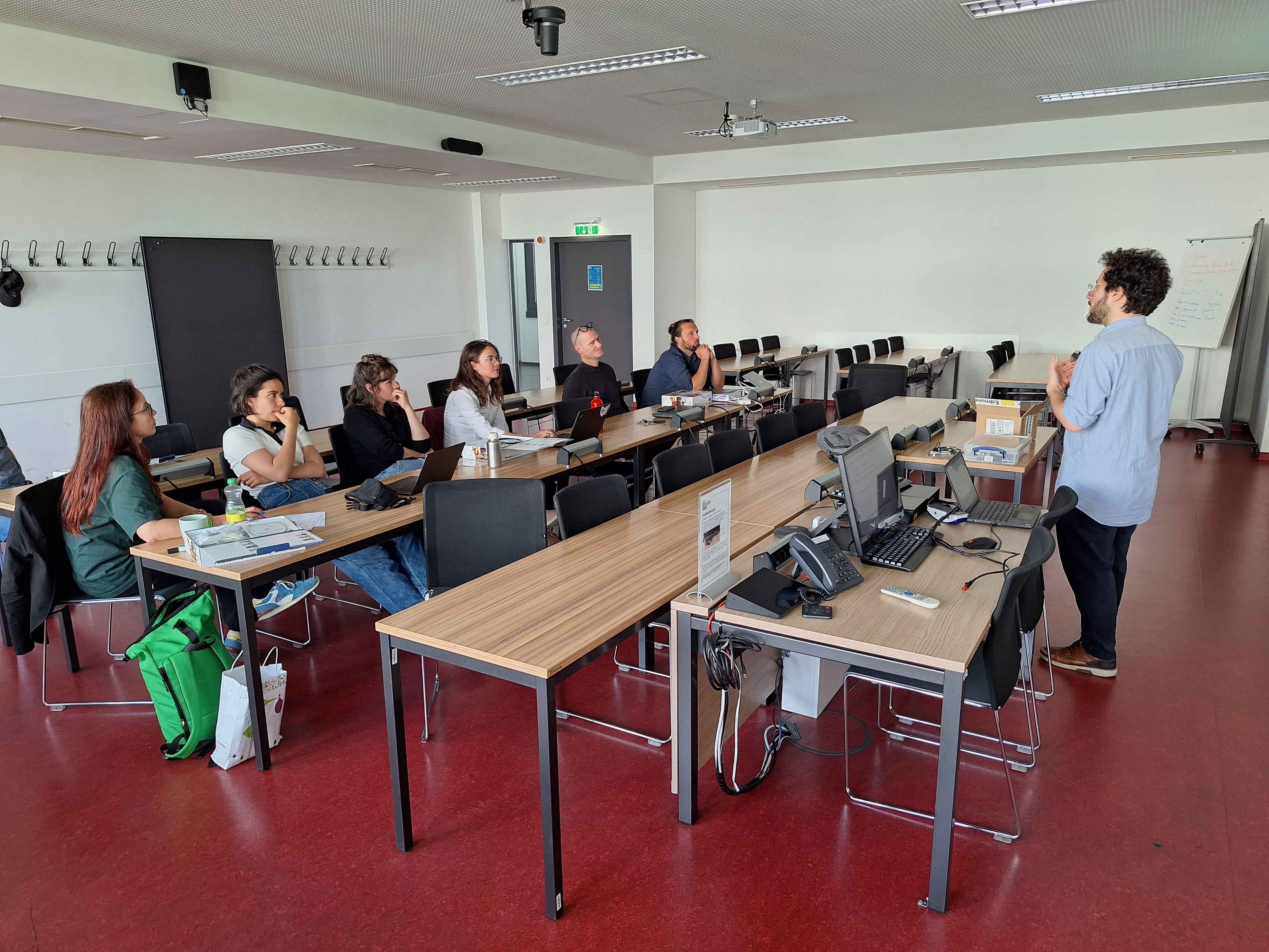 Ein Mann steht und spricht zu einer Gruppe von Menschen, die in einem Klassenzimmer an Schreibtischen sitzen. Auf den Tischen liegen Laptops, Papiere und Taschen. Der Raum hat einen roten Boden, weiße Wände und im Hintergrund eine Whiteboard-Tafel.