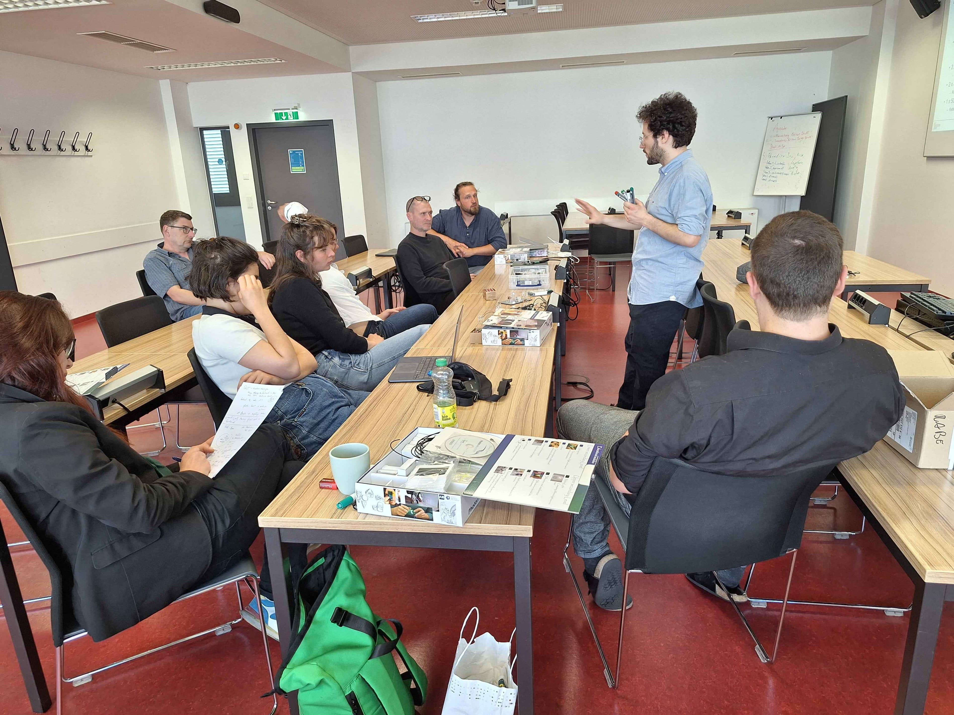 Ein Mann steht und spricht zu einer Gruppe von Menschen, die an Tischen in einem Klassenzimmer oder Besprechungsraum sitzen. Auf den Tischen liegen Papiere, Getränke und Taschen, und im Hintergrund ist eine Whiteboard-Tafel zu sehen.