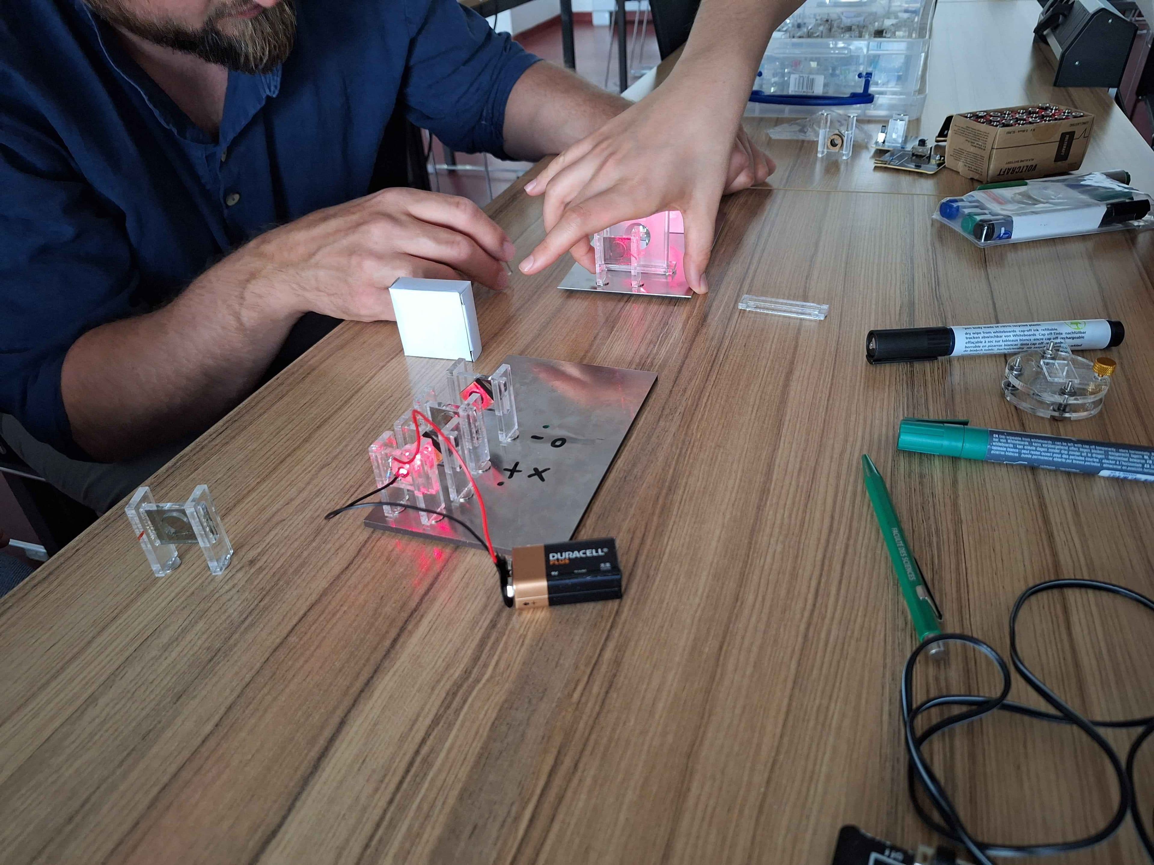 Zwei Personen führen ein wissenschaftliches Experiment auf einem Tisch durch, bei dem sie LED-Leuchten, durchsichtige Kunststoffteile, eine Metallplatte, Kabel, eine 9-V-Batterie sowie verschiedene kleine Werkzeuge und Marker verwenden.