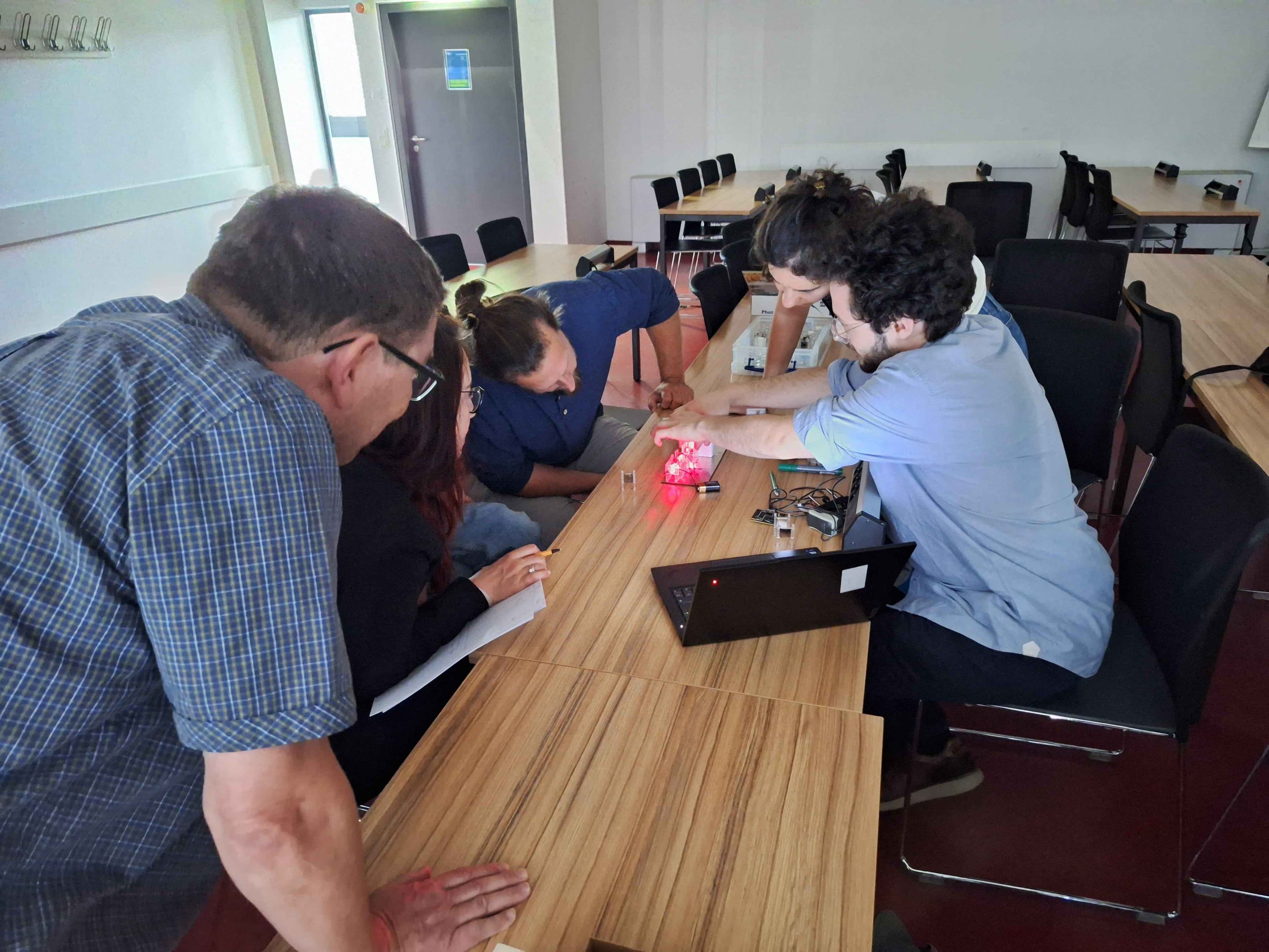 Vier Personen sitzen in einem Klassenzimmer um einen Holztisch herum und arbeiten konzentriert an einem Elektronikprojekt mit rot leuchtenden Lämpchen, während daneben ein Laptop steht.