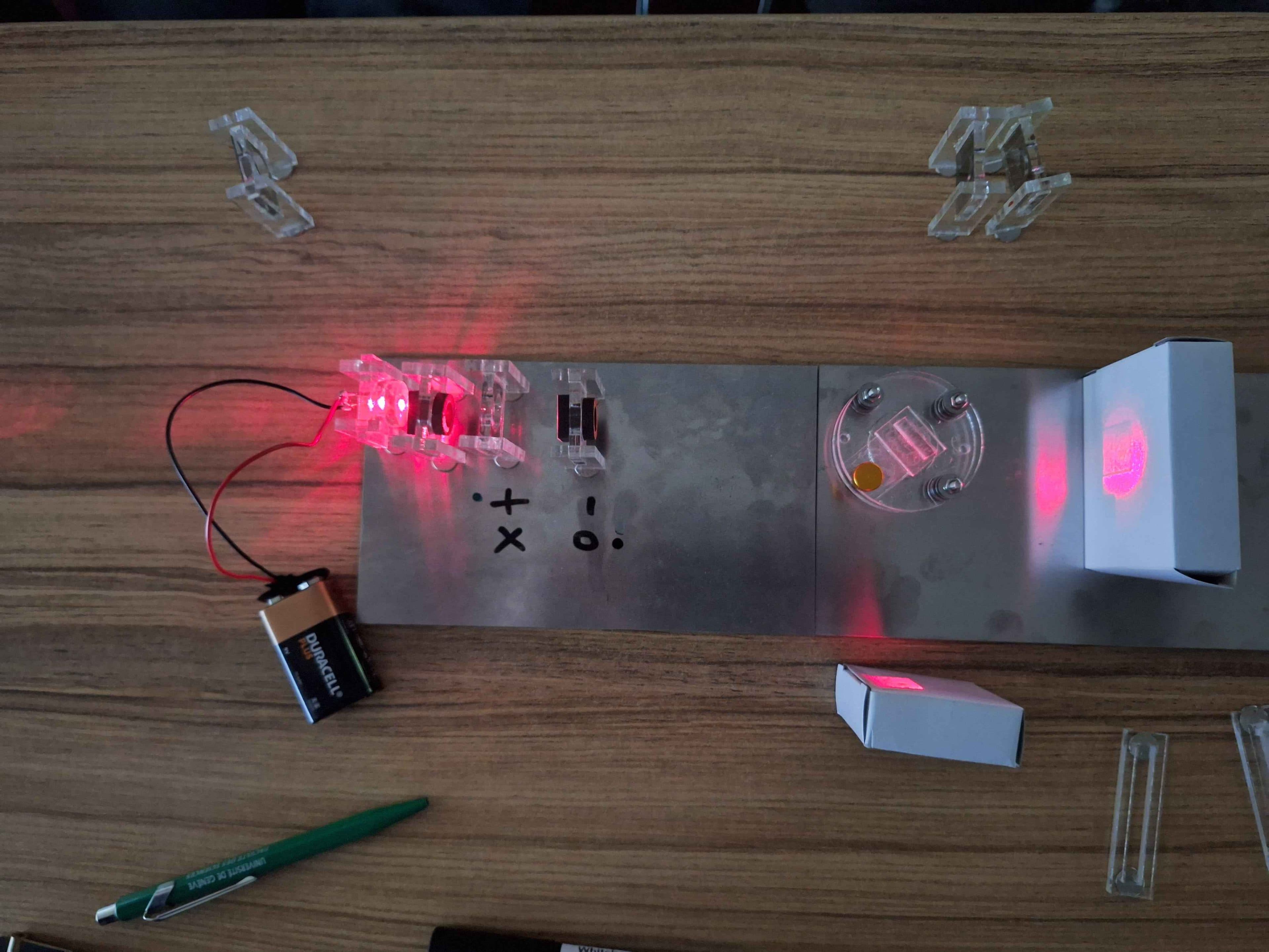 Ein wissenschaftliches Experiment auf einem Tisch mit einer Batterie, einer LED-Leuchte, Spiegeln und Glaselementen, die rotes Licht reflektieren, sowie mathematischen Symbolen auf einer Metallplatte. Ein grüner Stift liegt daneben auf der Holzoberfläche.