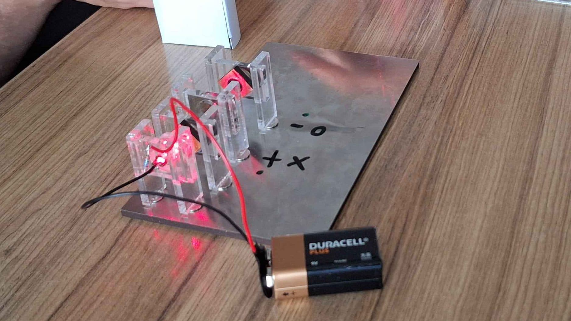 Zwei Personen arbeiten an einem Tisch an einem wissenschaftlichen Experiment mit einer Batterie, LEDs, Kabeln und durchsichtigen Kunststoffteilen. Eine Person justiert ein Teil, während die andere ihr dabei hilft. Auf dem Tisch liegen außerdem Stifte und Werkzeuge.