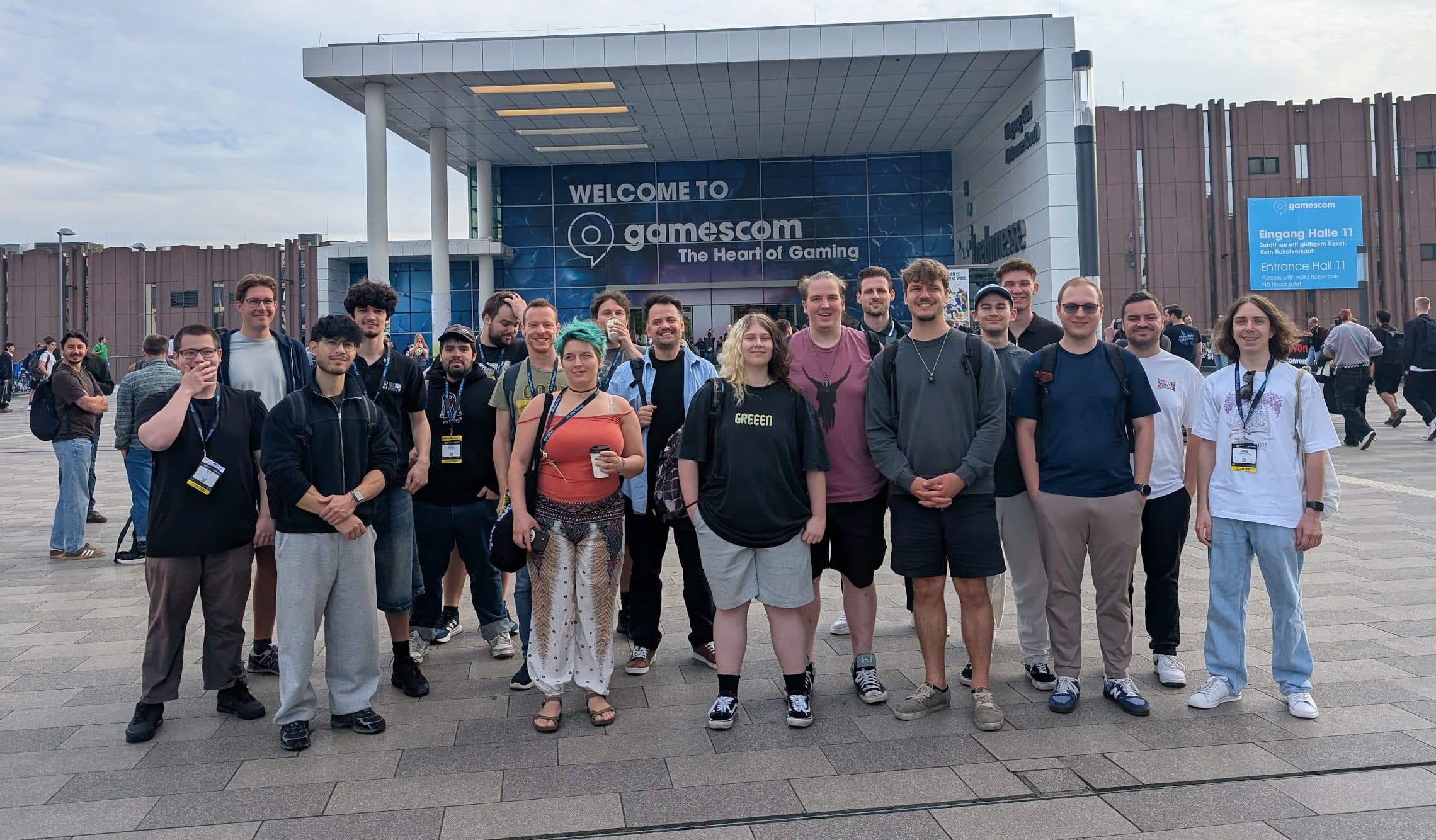 Eine Gruppe von Menschen steht vor einem großen Veranstaltungsort, über dessen Eingang ein Schild mit der Aufschrift Welcome to gamescom hängt, und lächelt für ein Foto. Im Hintergrund sind weitere Besucher und das Gebäude zu sehen.