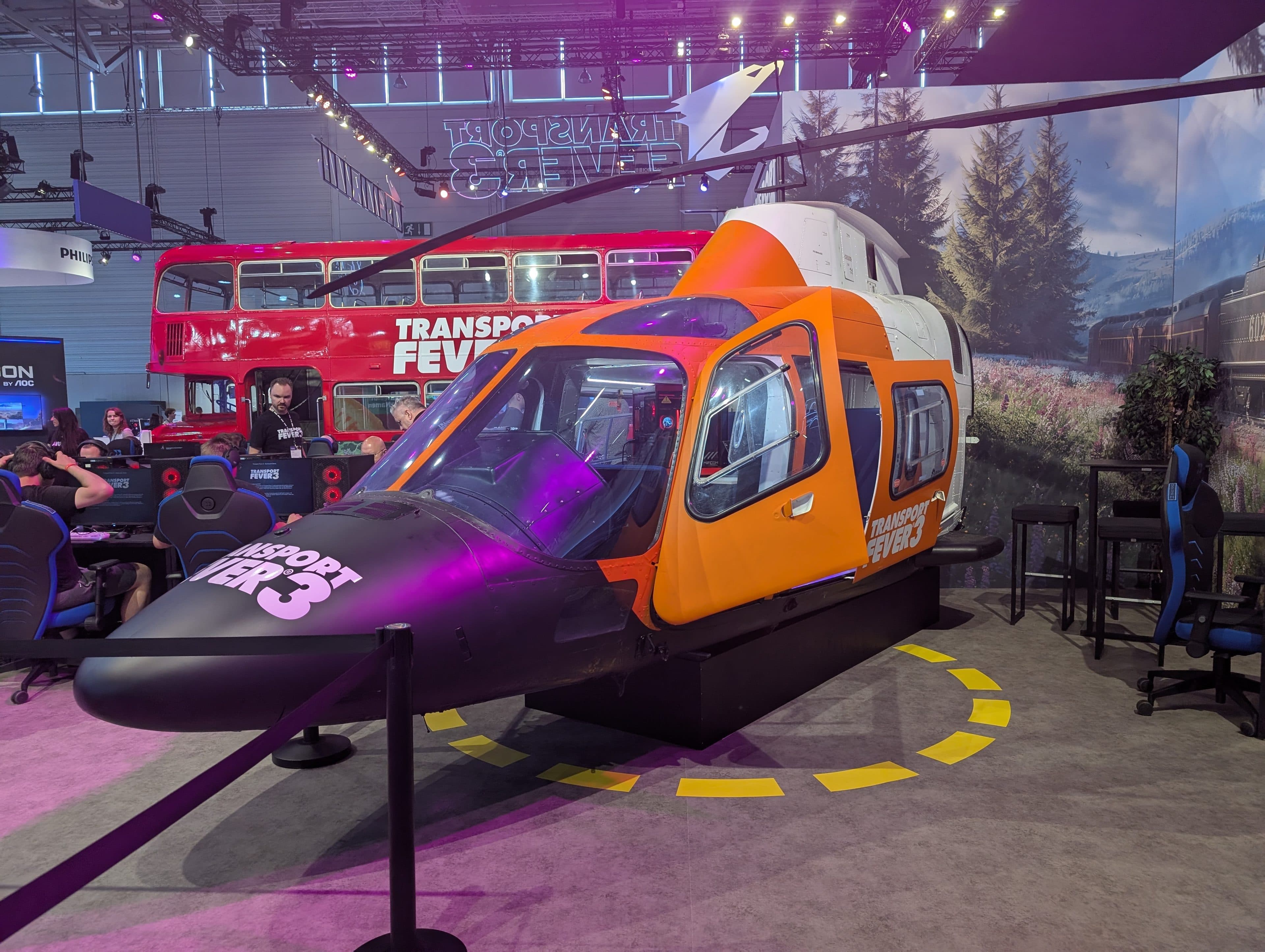 Im Innenbereich ist ein orange-weißer Hubschraubersimulator in Originalgröße für Transport Fever 3 ausgestellt, mit Menschen in der Nähe und einem roten Doppeldeckerbus im Hintergrund.