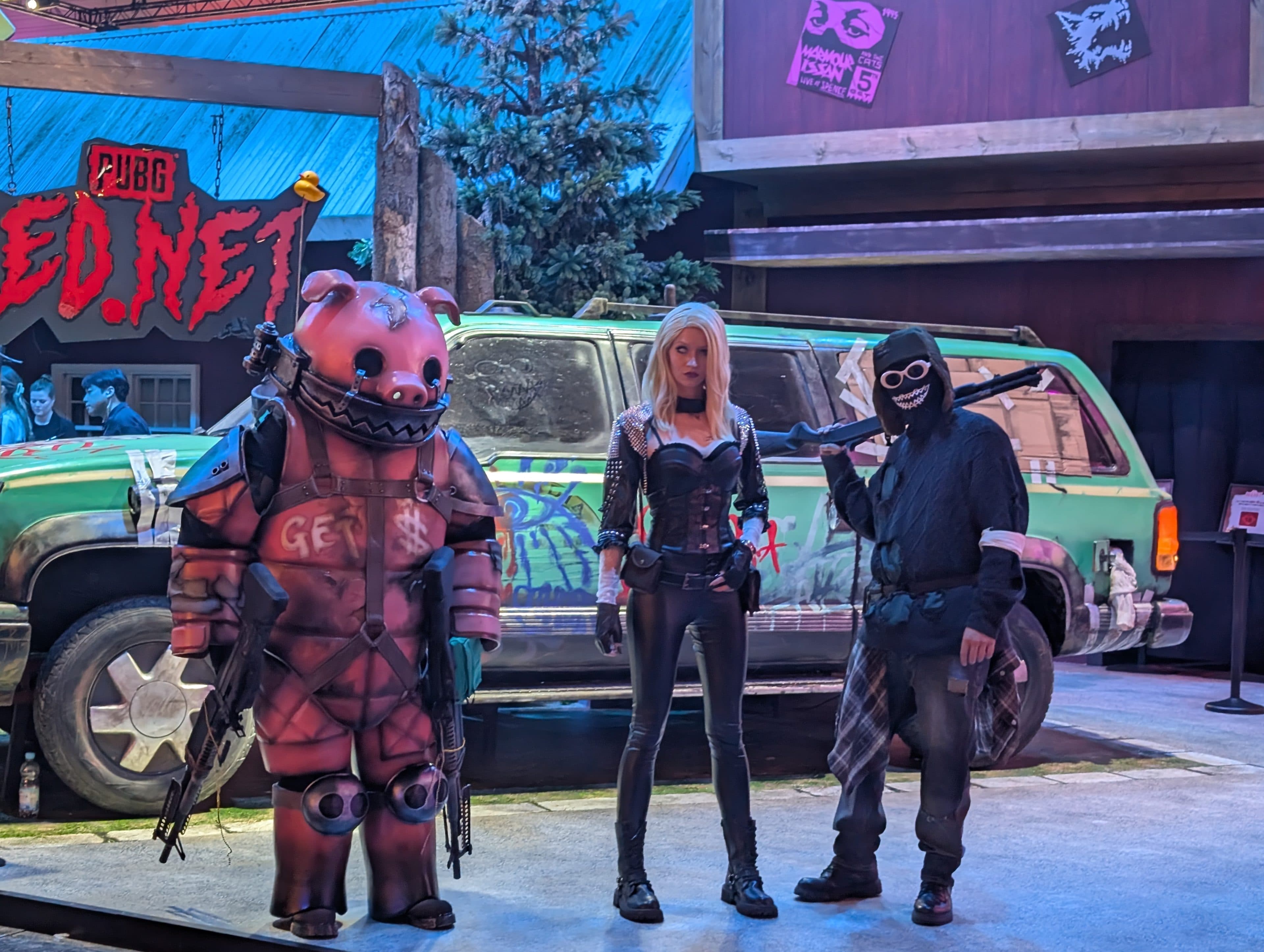 Drei Personen in Cosplay-Kostümen stehen vor einem grünen, mit Graffiti besprühten Fahrzeug; eine trägt eine rosa Schweinemaske, eine andere eine blonde Perücke und einen schwarzen Anzug, und die dritte eine Kapuze mit Schutzbrille und hält einen Schläger in der Hand. Die Kulisse wirkt postapokalyptisch.
