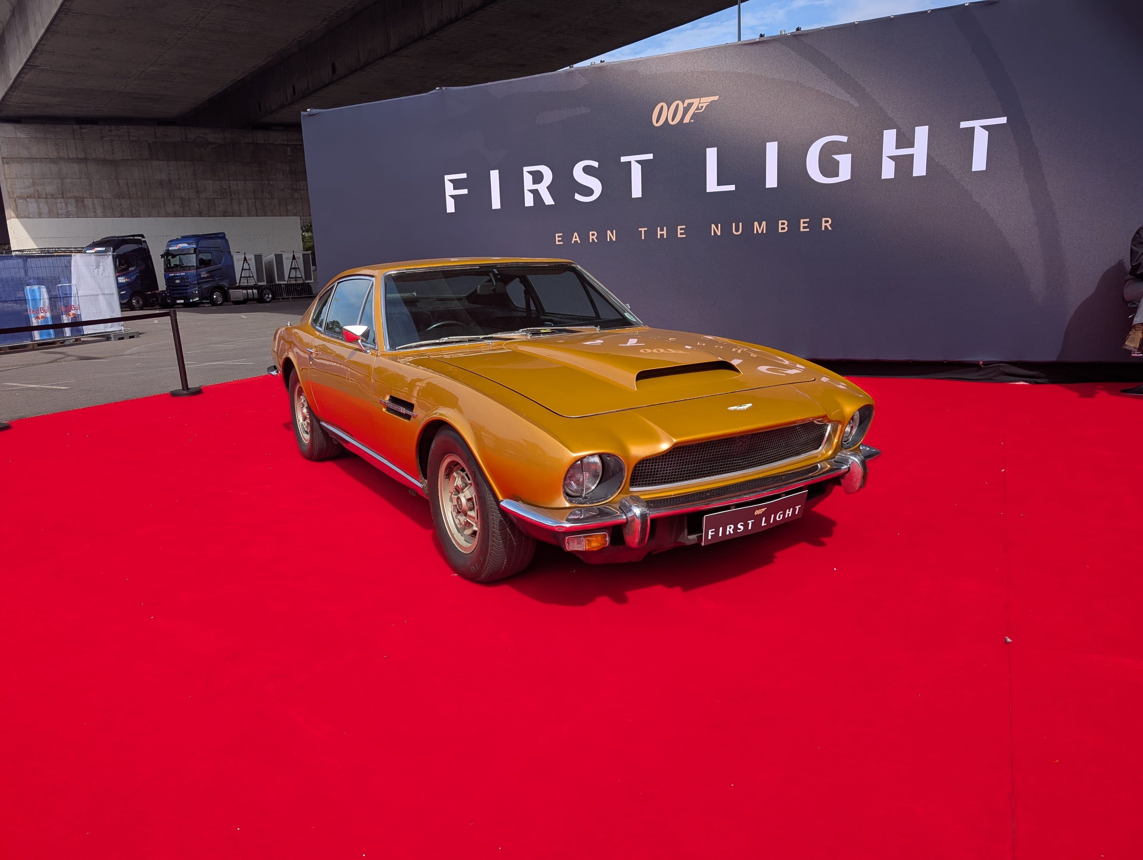 Ein klassisches goldenes Auto auf einem roten Teppich vor einem Banner mit der Aufschrift 007 First Light: Earn the Number, das bei einer Veranstaltung im Freien unter einer Überführung zu sehen ist.