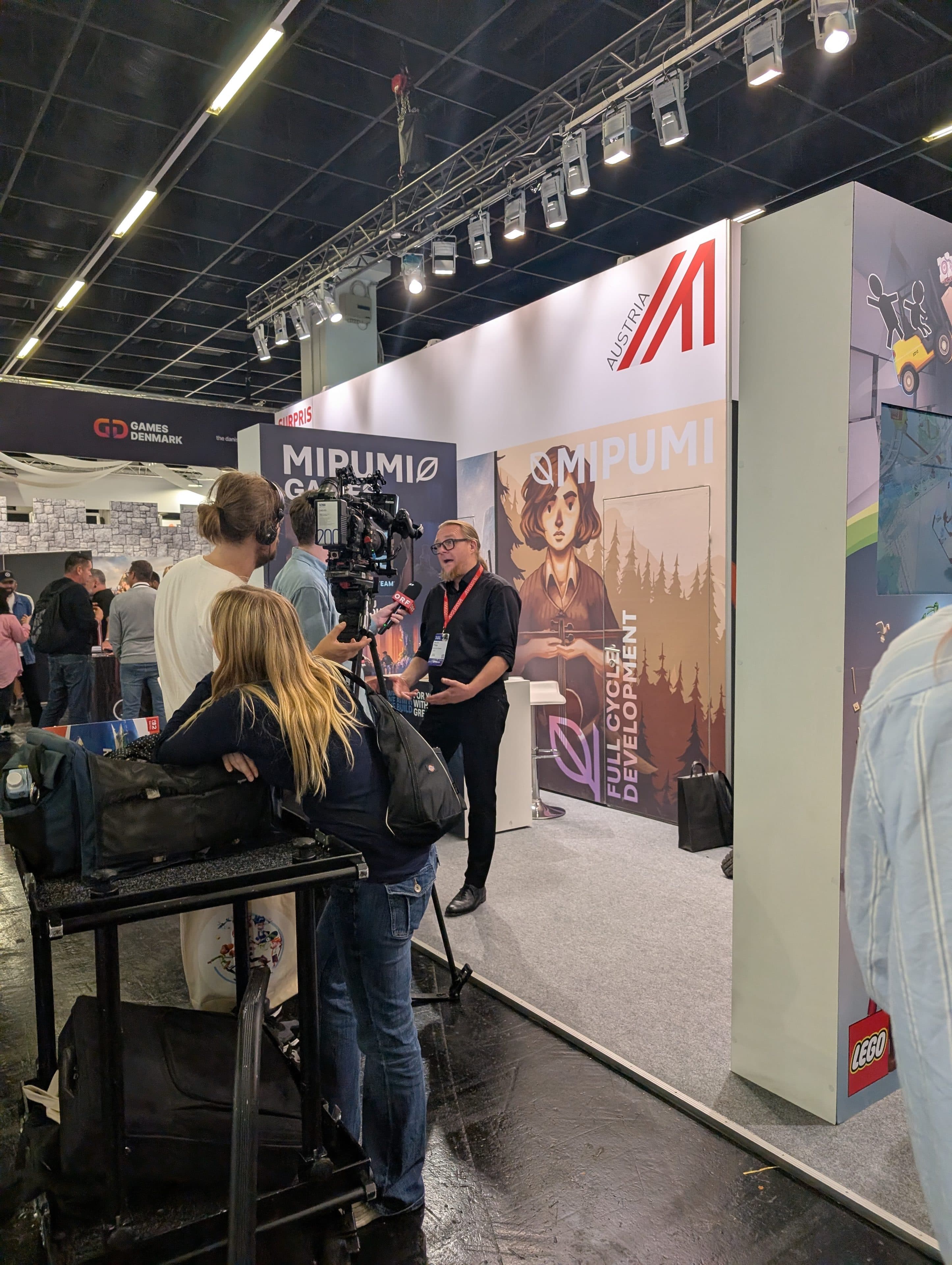 Ein Mann wird von einem Kamerateam am Stand von Mipumi Games auf einer Messe gefilmt. Besucher schauen zu, und es gibt Banner, Beleuchtung und Ausstellungsstücke, darunter ein deutlich sichtbares LEGO-Logo an der Ecke des Standes.