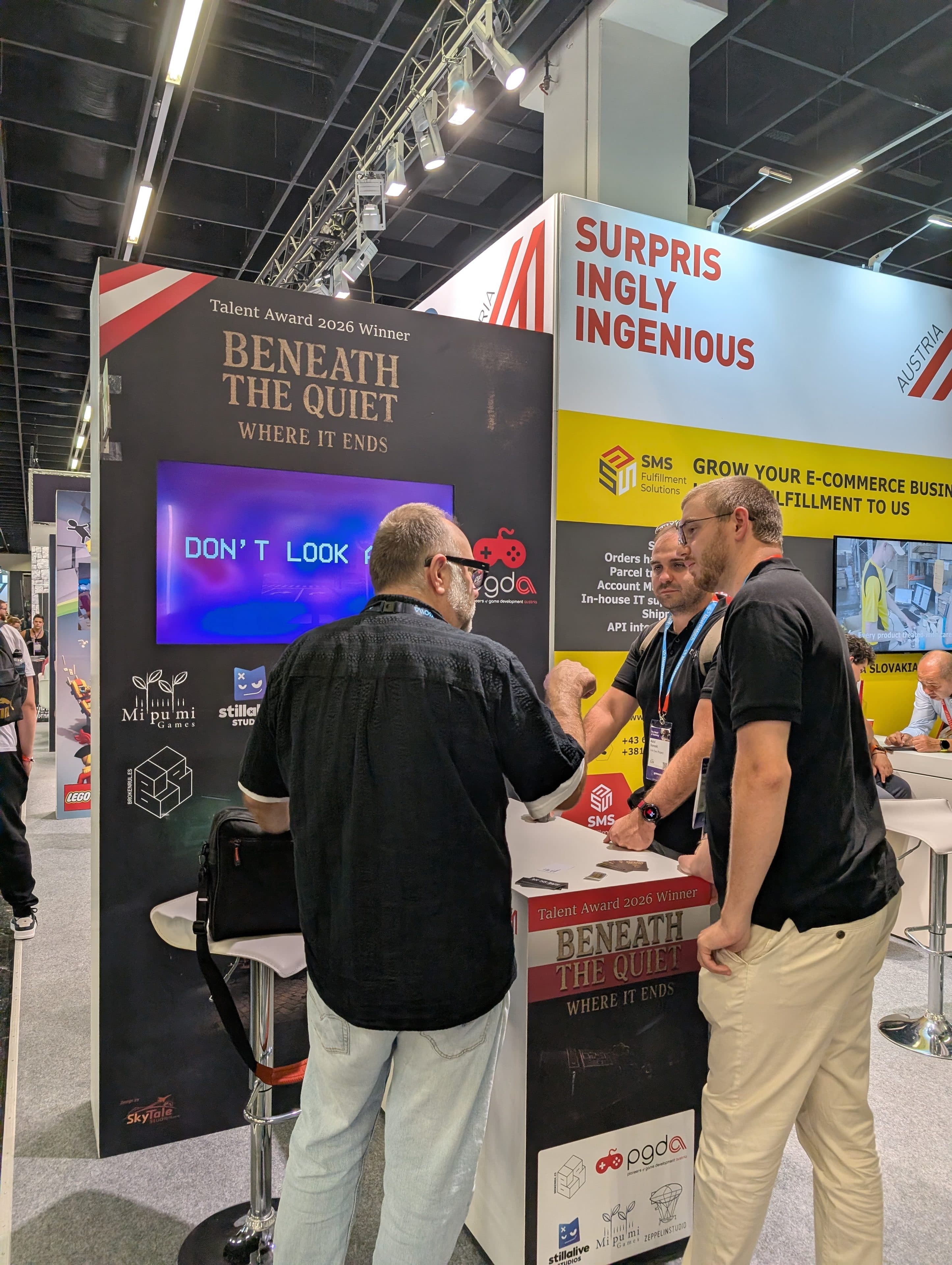 Drei Männer stehen vor einem Messestand auf einer Messe und unterhalten sich. Am Stand ist ein Schild mit der Aufschrift Beneath the Quiet zu sehen sowie eine Leinwand mit der Aufschrift DONT LOOK. Im Hintergrund sind bunte Banner und Messestände zu erkennen.