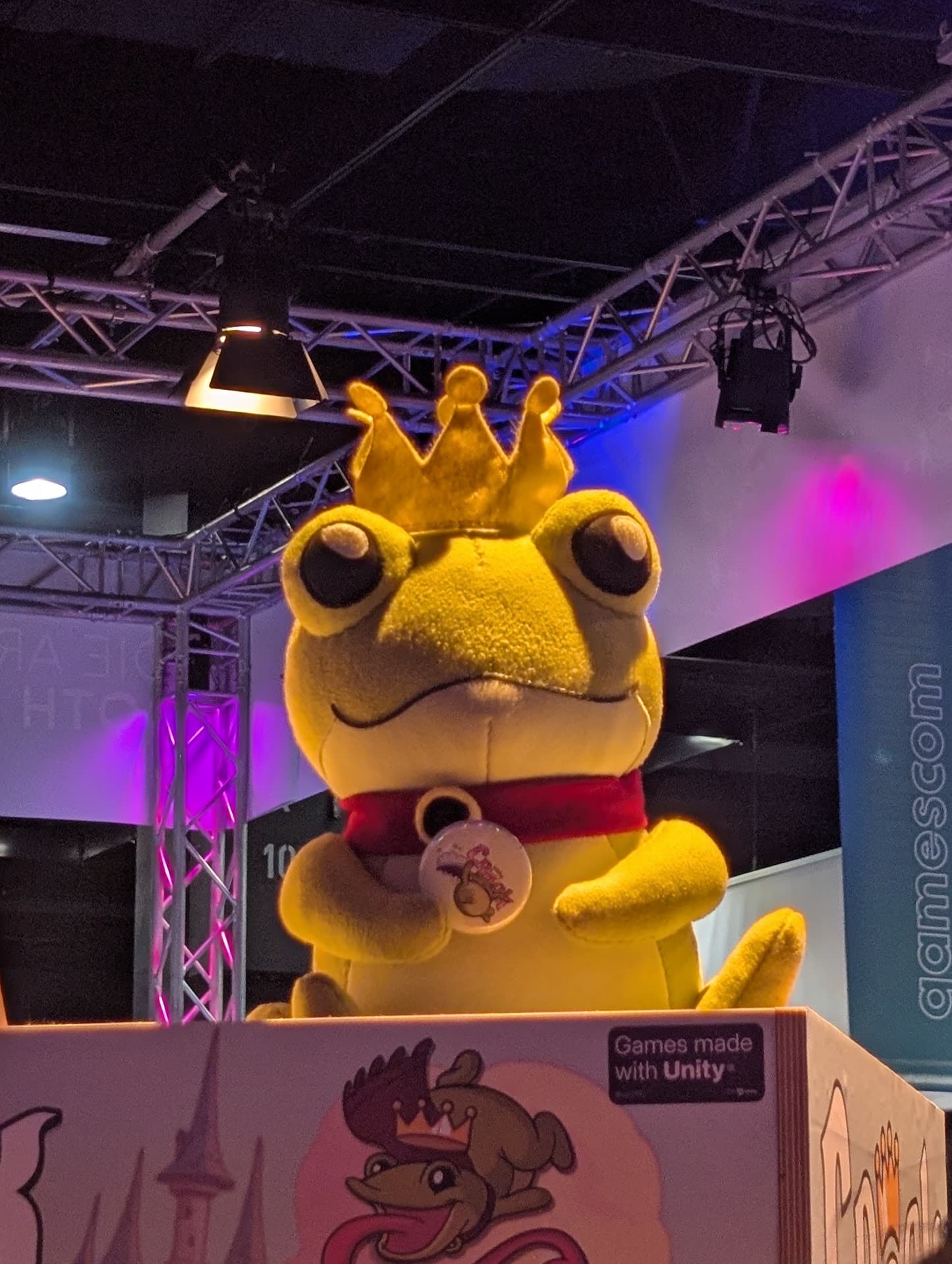 An einem Stand auf einer Veranstaltung ist ein großer gelber Plüschfrosch mit einer goldenen Krone und einem roten Umhang ausgestellt; daneben befinden sich bunte Lichter und ein Schild mit der Aufschrift Games made with Unity.