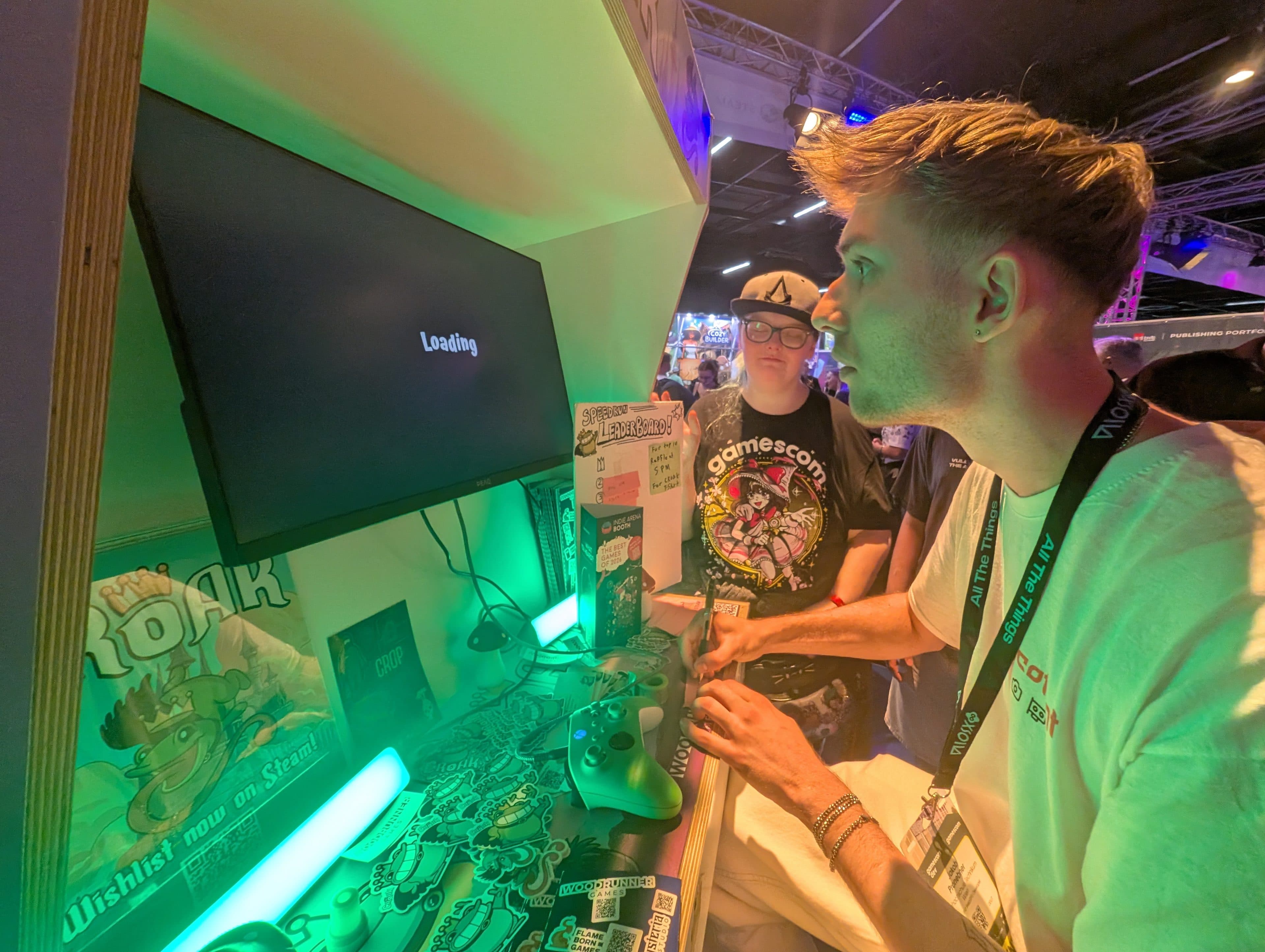 Eine Person sitzt auf einer Gaming-Veranstaltung an einem Stand und spielt ein Spiel, während auf dem Bildschirm Loading angezeigt wird. Hinter ihr steht eine weitere Person, und auf dem Tisch liegen verschiedene Gaming-Fanartikel und Dekorationen.