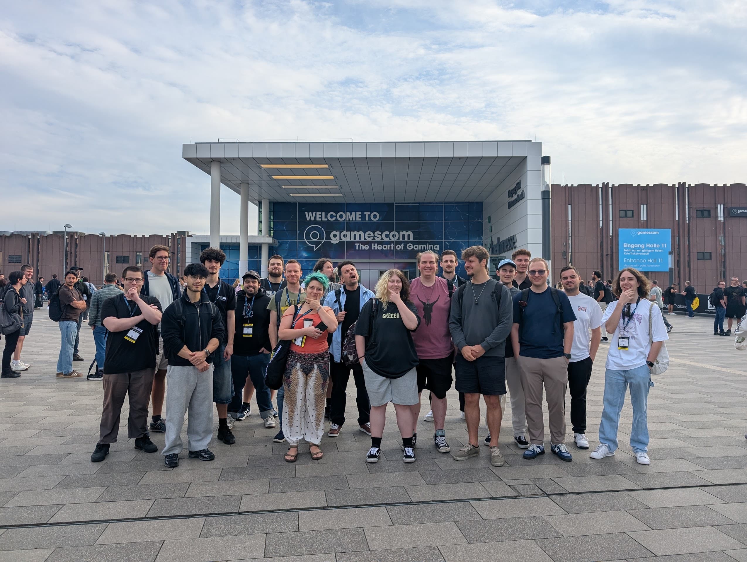 Eine Gruppe von Menschen posiert lächelnd vor dem Eingang der Gamescom; hinter ihnen ist unter einem bewölkten Himmel das Veranstaltungslogo am Gebäude zu sehen.