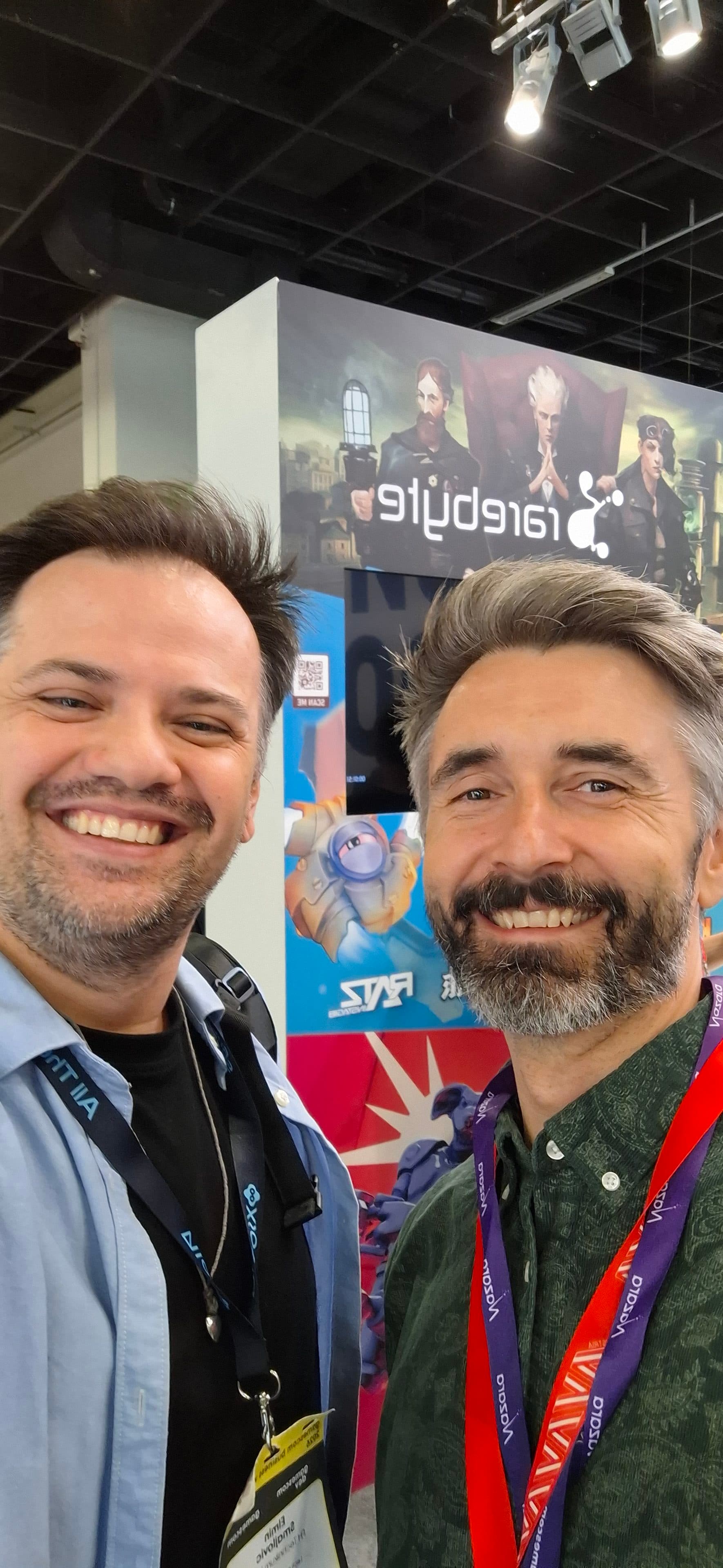 Zwei lächelnde Männer machen bei einer Veranstaltung ein Selfie, während sie vor einem bunten Stand stehen, hinter dem auf einem großen Plakat das Traplight-Logo und Spielfiguren zu sehen sind.