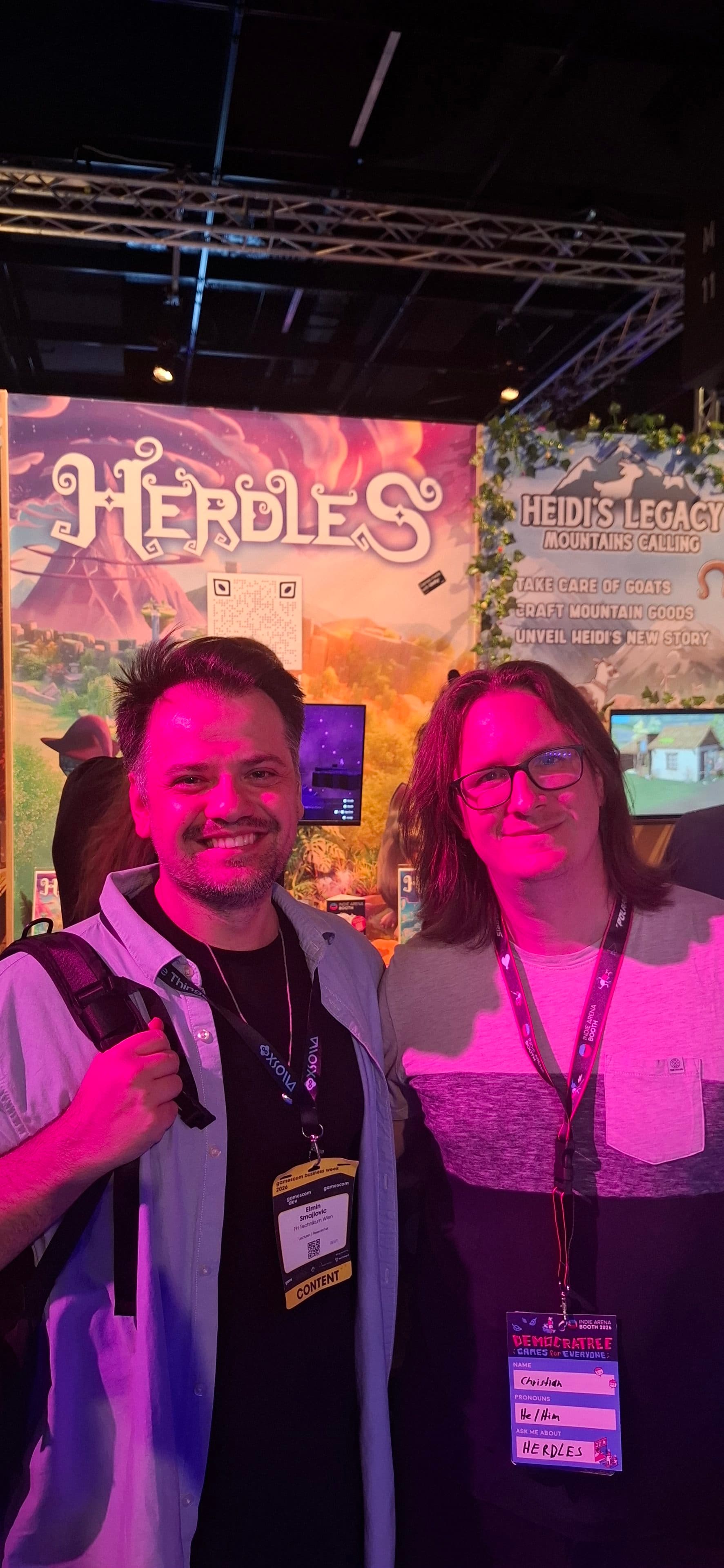 Zwei Männer stehen lächelnd vor einem bunten Herdles-Spielestand auf der Gamescom. Der Mann links hat kurzes dunkles Haar und trägt ein hellblaues Hemd; der Mann rechts hat langes dunkles Haar, eine Brille und ein graues Hemd. Beide tragen Messebadges.