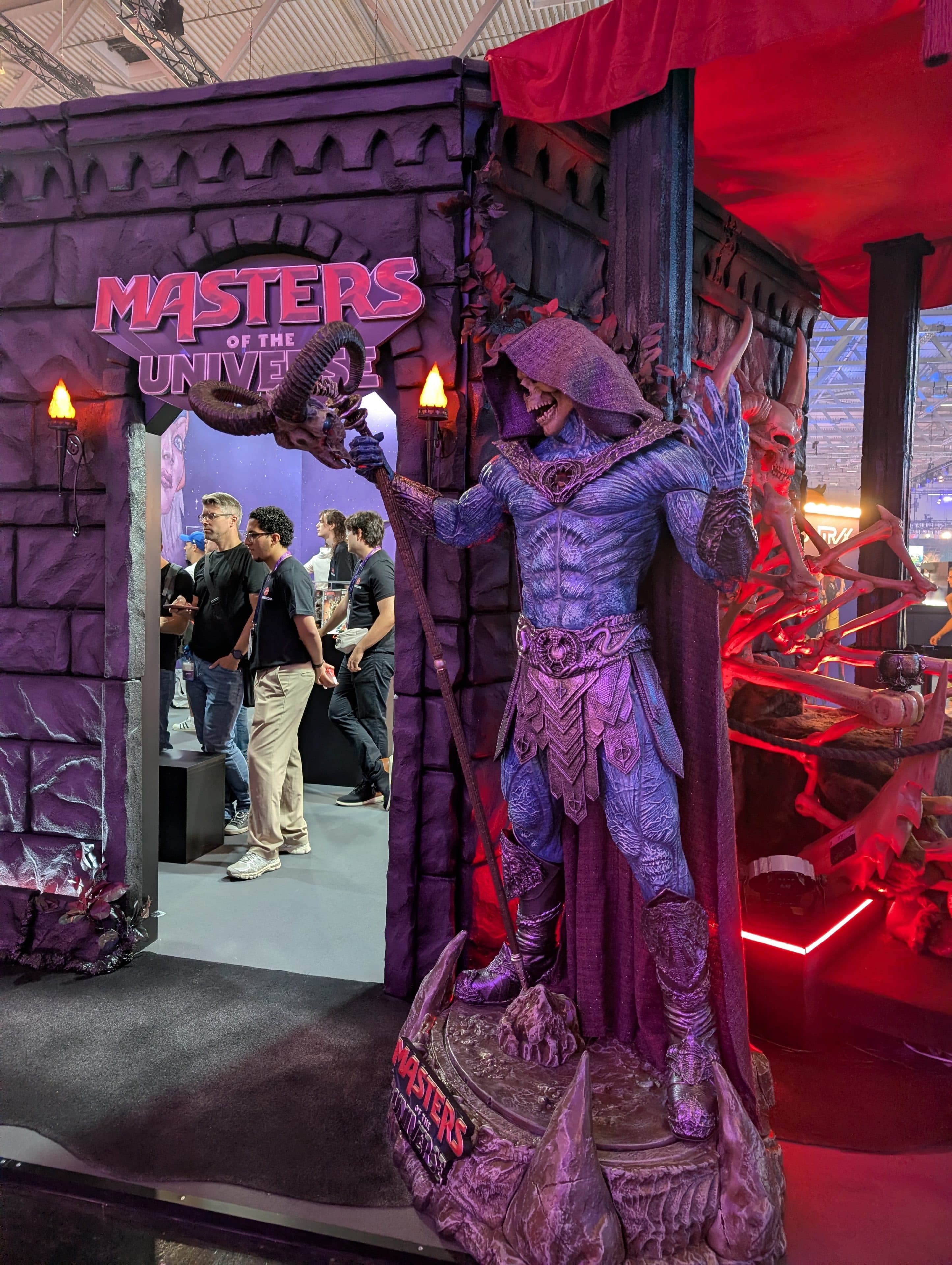 Am Eingang des Masters of the Universe-Standes steht eine lebensgroße Skeletor-Statue; Besucher betreten den Stand, der von violetten, burgartigen Wänden umgeben ist.
