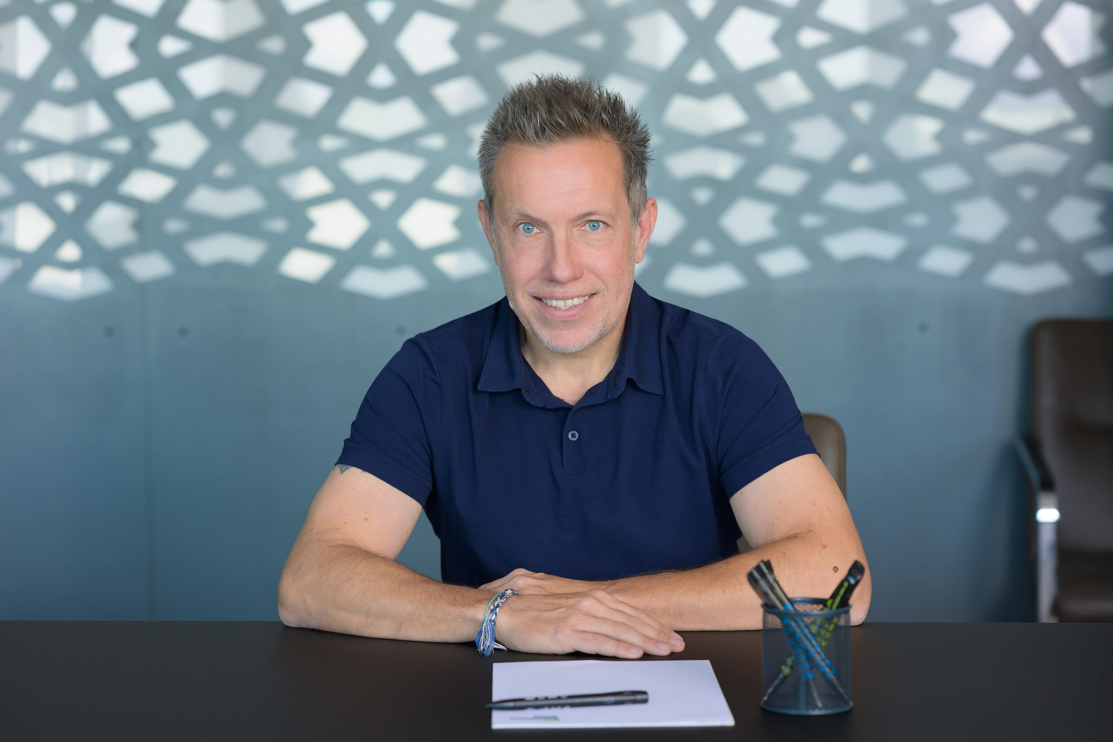 Ein Mann mit kurzen grauen Haaren in einem marineblauen Polohemd sitzt an einem Schreibtisch mit einem Notizblock und einem Stifthalter und lächelt in die Kamera. Im Hintergrund sind eine gemusterte Wand und ein leerer Stuhl zu sehen.