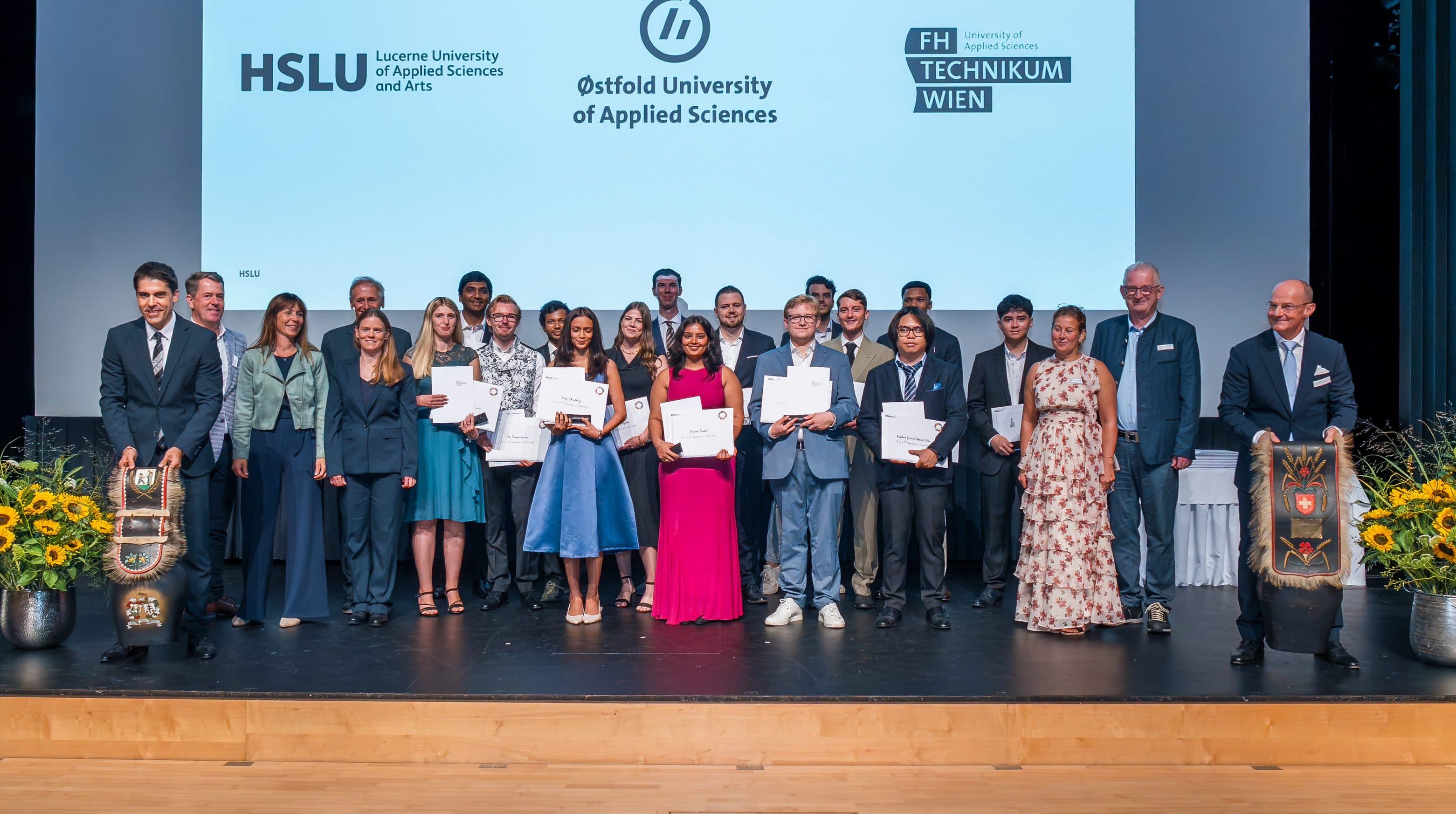Eine Gruppe von Absolventen und Dozenten posiert auf der Bühne mit ihren Diplomen und lächelt vor einer Leinwand, auf der Herzlichen Glückwunsch zum Abschluss! sowie die Logos der Universität zu lesen sind. Blumen und Dekorationen säumen die Bühne.