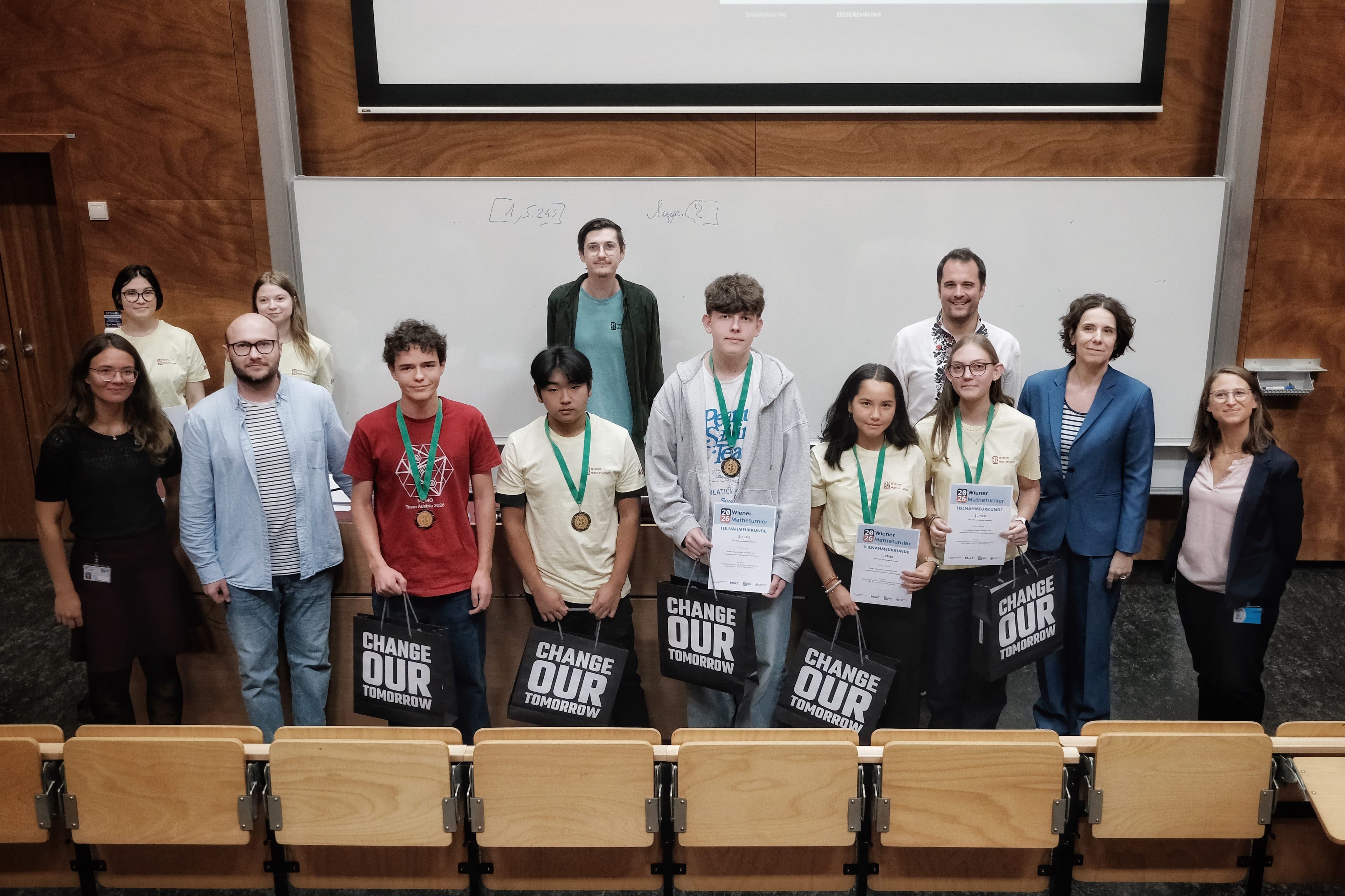 Eine Gruppe von Schülern, die Medaillen tragen und Urkunden in den Händen halten, steht vor einer Tafel, hinter ihnen befinden sich Erwachsene in einem Hörsaal. Einige Schüler halten Taschen mit der Aufschrift Change Our Tomorrow in den Händen.