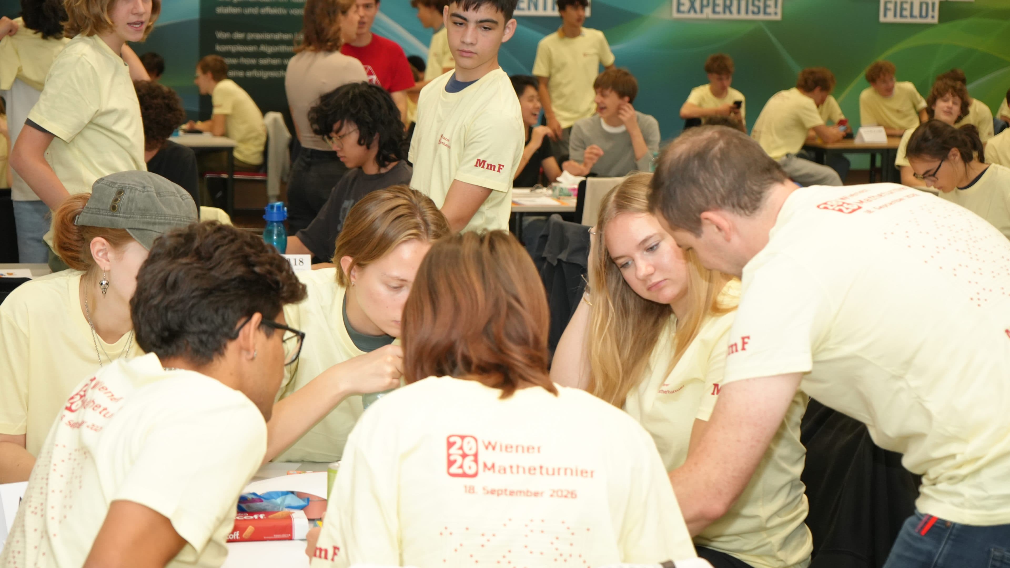 Eine Gruppe von Schülern in einheitlichen T-Shirts arbeitet während eines Mathematikwettbewerbs gemeinsam an einem Tisch, während im Hintergrund weitere Schüler in einem großen, hell beleuchteten Raum arbeiten.