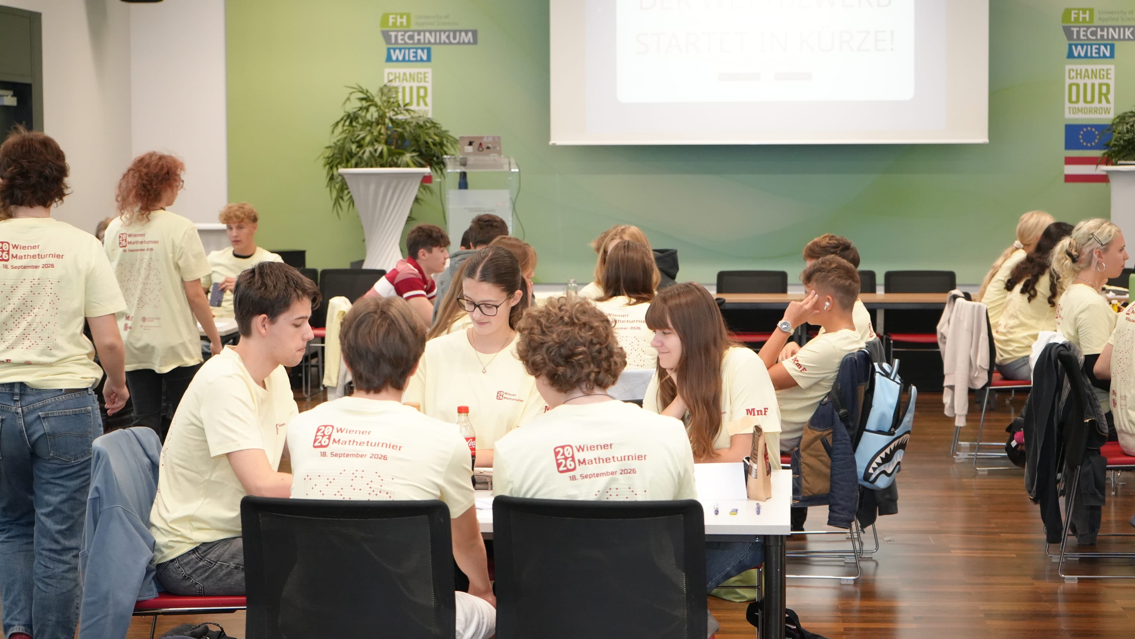 Eine Gruppe junger Menschen in einheitlichen T-Shirts sitzt an Tischen in einem hellen Raum und unterhält sich, während sie sich auf eine Veranstaltung vorbereitet. Im Hintergrund sind eine Projektionswand und eine grüne Wand mit Motivationspostern zu sehen.