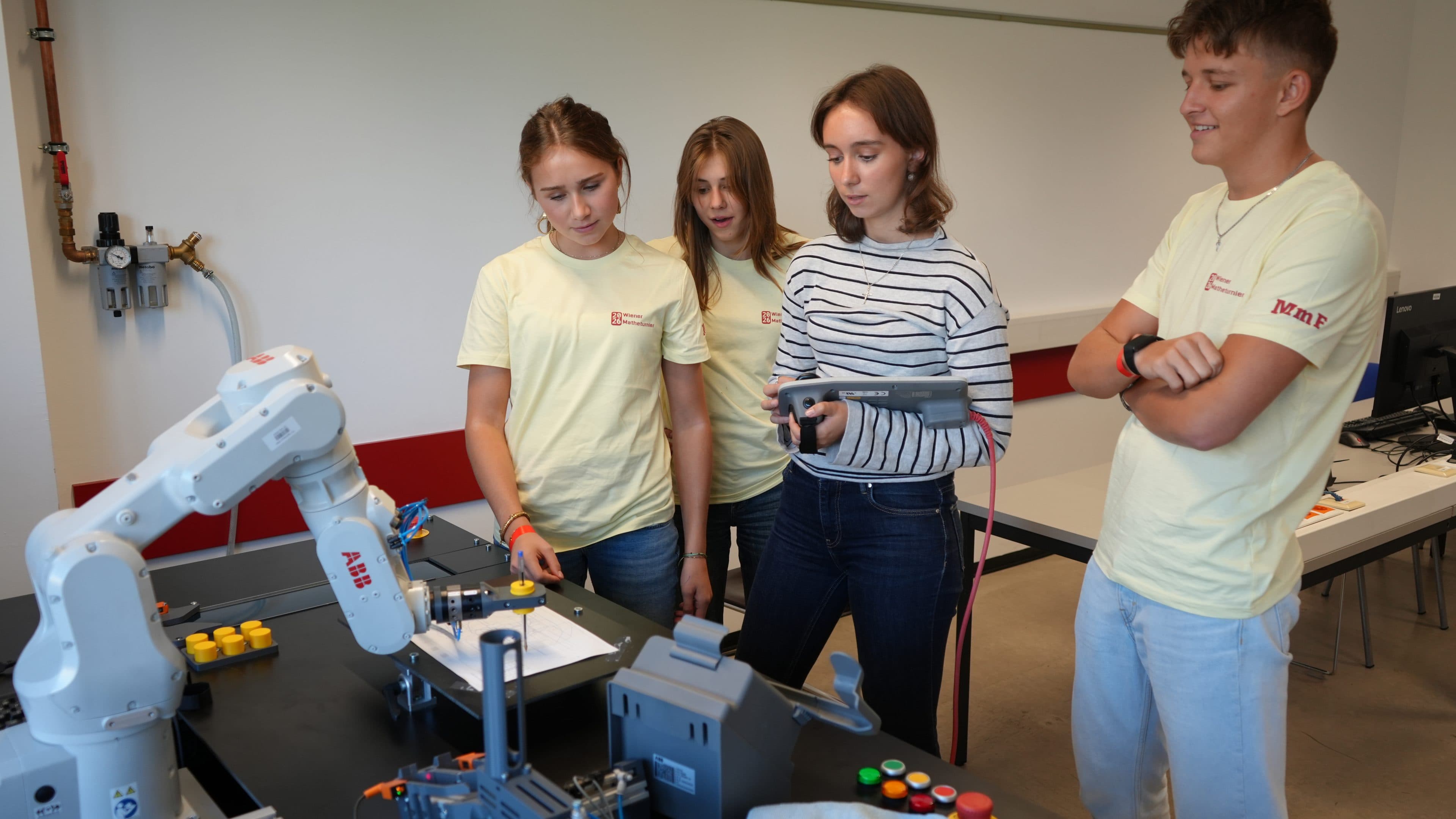 Vier junge Leute beobachten und bedienen an einem Tisch einen Roboterarm; zwei von ihnen tragen gelbe Hemden, einer hält einen Controller in der Hand. Im Hintergrund sind Laptops und Geräte zu sehen.