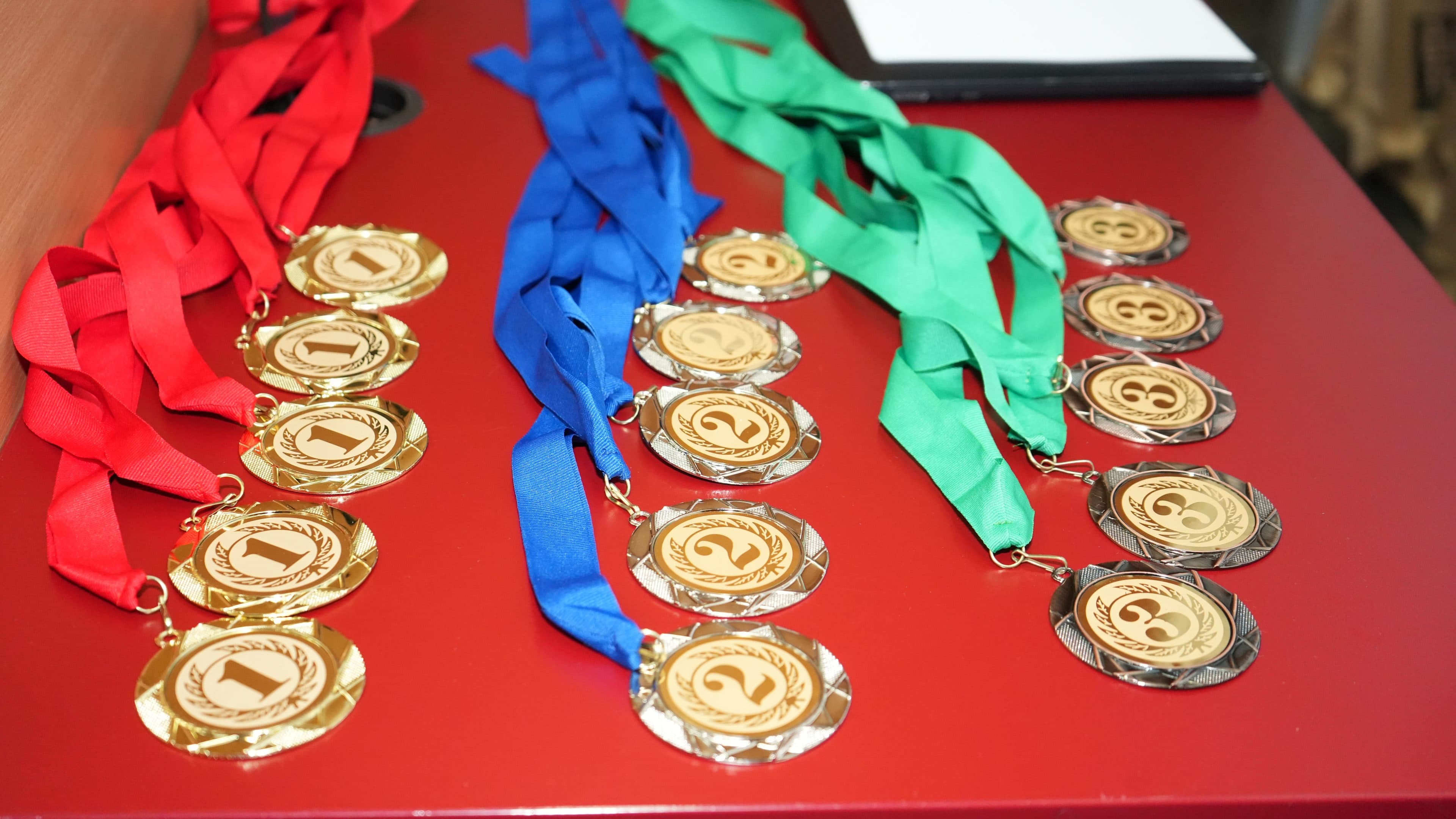 Auf einem roten Tisch liegen Goldmedaillen mit roten, blauen und grünen Bändern. Auf jeder Medaille ist eine Zahl zwischen 1 und 3 zu sehen. Außerdem befindet sich in der oberen rechten Ecke des Bildes ein weißes, rechteckiges Objekt.
