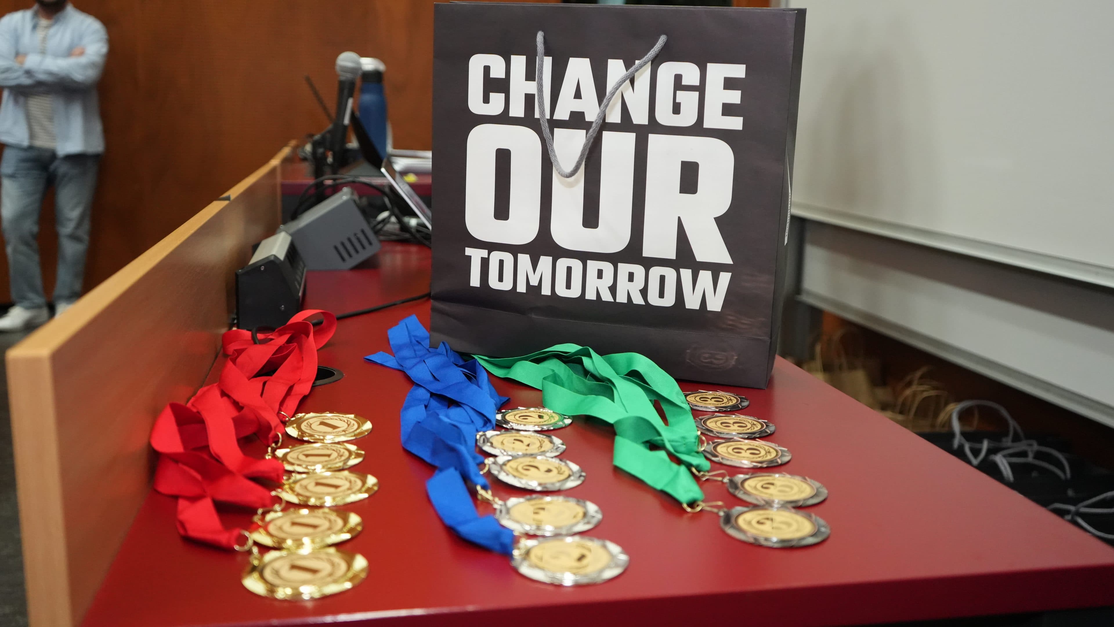 Auf einem roten Tisch neben einer Tasche mit der Aufschrift Change Our Tomorrow liegen Medaillen mit roten, blauen und grünen Bändern. Im Hintergrund steht ein Mann.