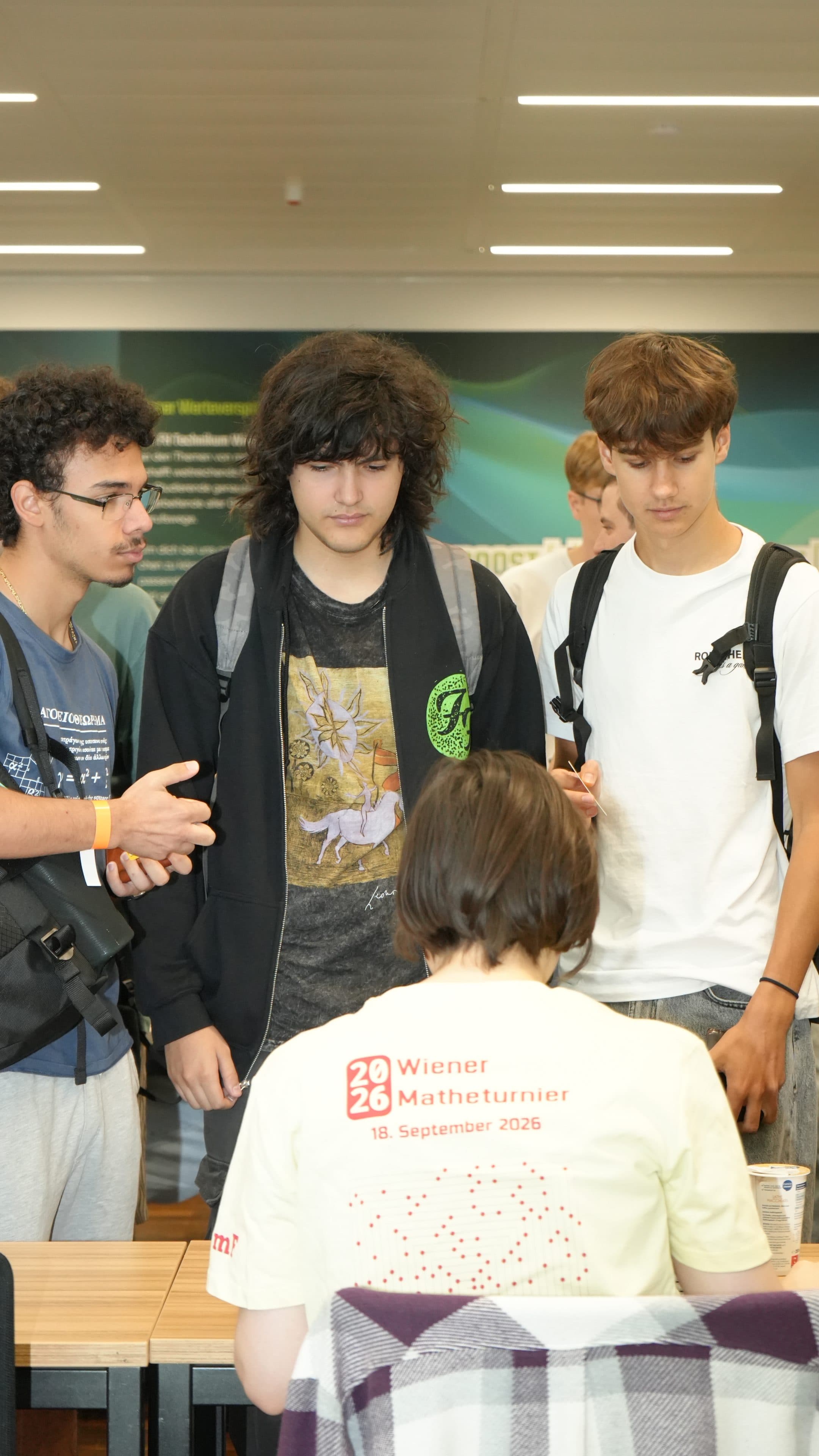 Drei junge Männer mit Rucksäcken stehen da und unterhalten sich mit einer Person, die an einem Schreibtisch sitzt und ein gelbes T-Shirt mit der Aufschrift Wiener Mathematikturnier 2026 trägt. Im Hintergrund ist eine moderne Innenraumkulisse zu sehen.