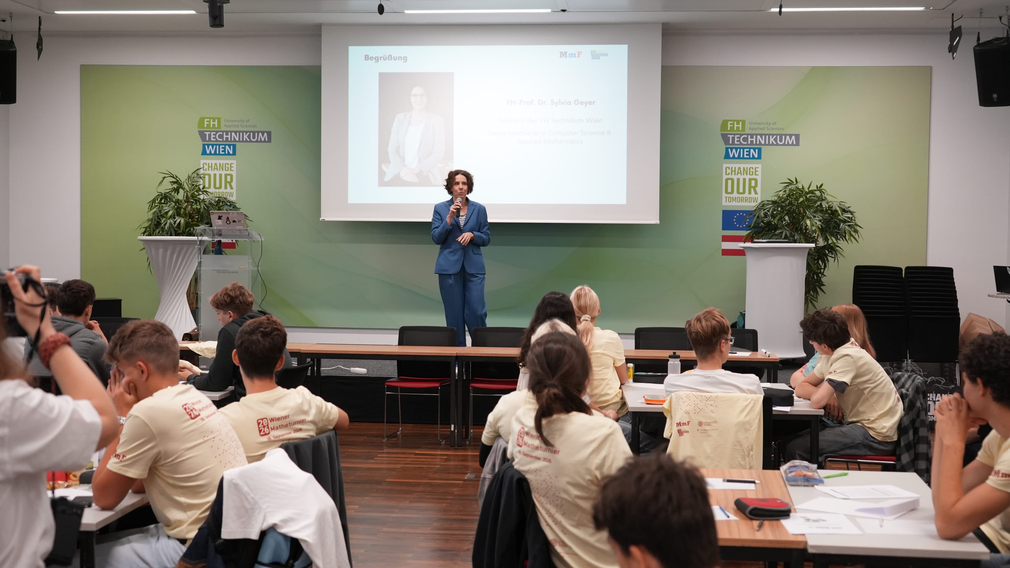 Eine Person in einem blauen Anzug spricht auf der Bühne zu einem Publikum, das an Tischen sitzt. Im Hintergrund sind eine Präsentationsfolie und die Aufschrift FH Technikum Wien zu sehen. Einige Teilnehmer tragen einheitliche beige Hemden.