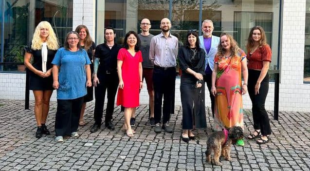 Eine Gruppe von zwölf Erwachsenen posiert lächelnd im Freien vor einem großen Gebäude mit vielen Fenstern, während im Vordergrund ein kleiner brauner Hund auf dem Kopfsteinpflaster steht.