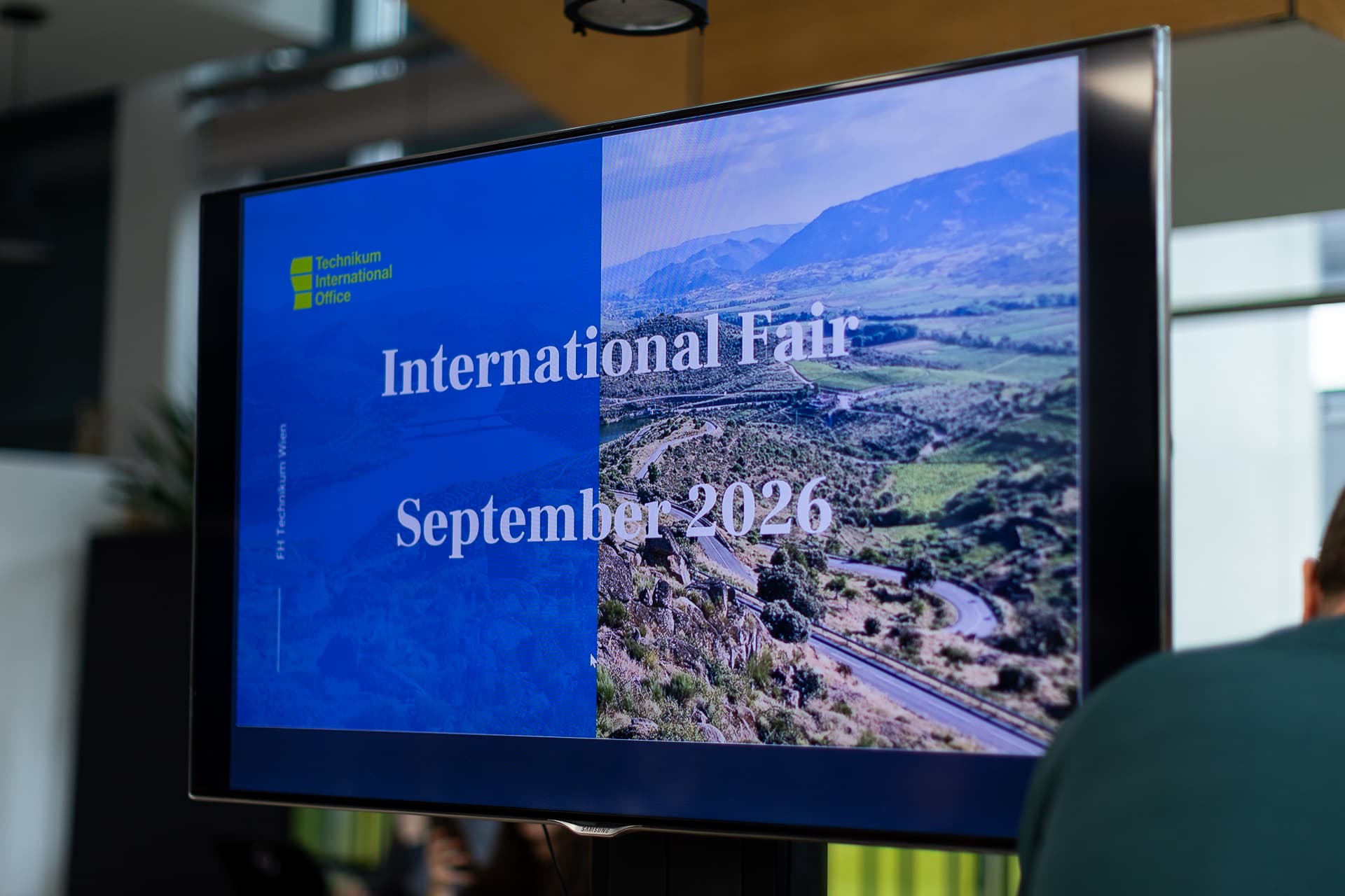 Auf einem Bildschirm wird eine Ankündigung für eine internationale Messe im September 2026 angezeigt, mit einem Landschaftsfoto und dem Logo des International Office des Technikums auf der linken Seite.