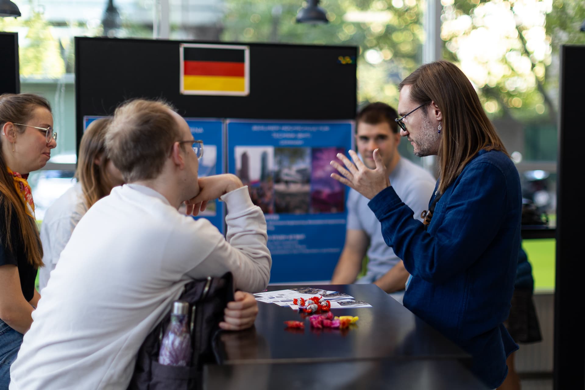 Eine Gruppe von Menschen unterhält sich an einem Tisch, auf dem Broschüren und Süßigkeiten liegen. Hinter ihnen sind Plakate und eine deutsche Flagge zu sehen. Im Hintergrund sind helle Fenster zu erkennen.