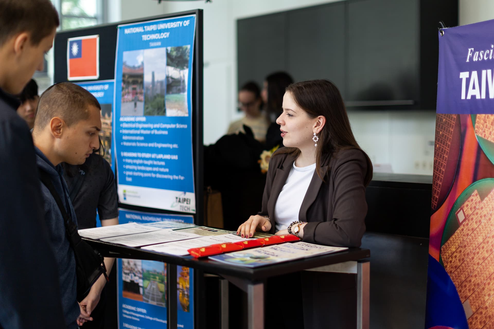 Eine Frau unterhält sich mit zwei Personen an einem Informationsstand, an dem Plakate über Taipeh und Taiwan ausgestellt sind. Auf dem Tisch liegen Broschüren und ein roter Gegenstand. Der Schauplatz scheint eine Bildungs- oder Kulturveranstaltung in einem Innenraum zu sein.