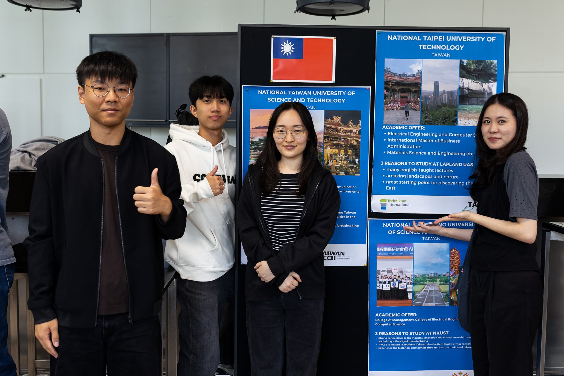 Vier Personen stehen neben zwei Informationsplakaten von Hochschulen – eines für die Nationale Taiwan-Universität für Wissenschaft und Technologie und eines für die Nationale Technische Universität Taipeh – und posieren. Eine Person streckt den Daumen nach oben.