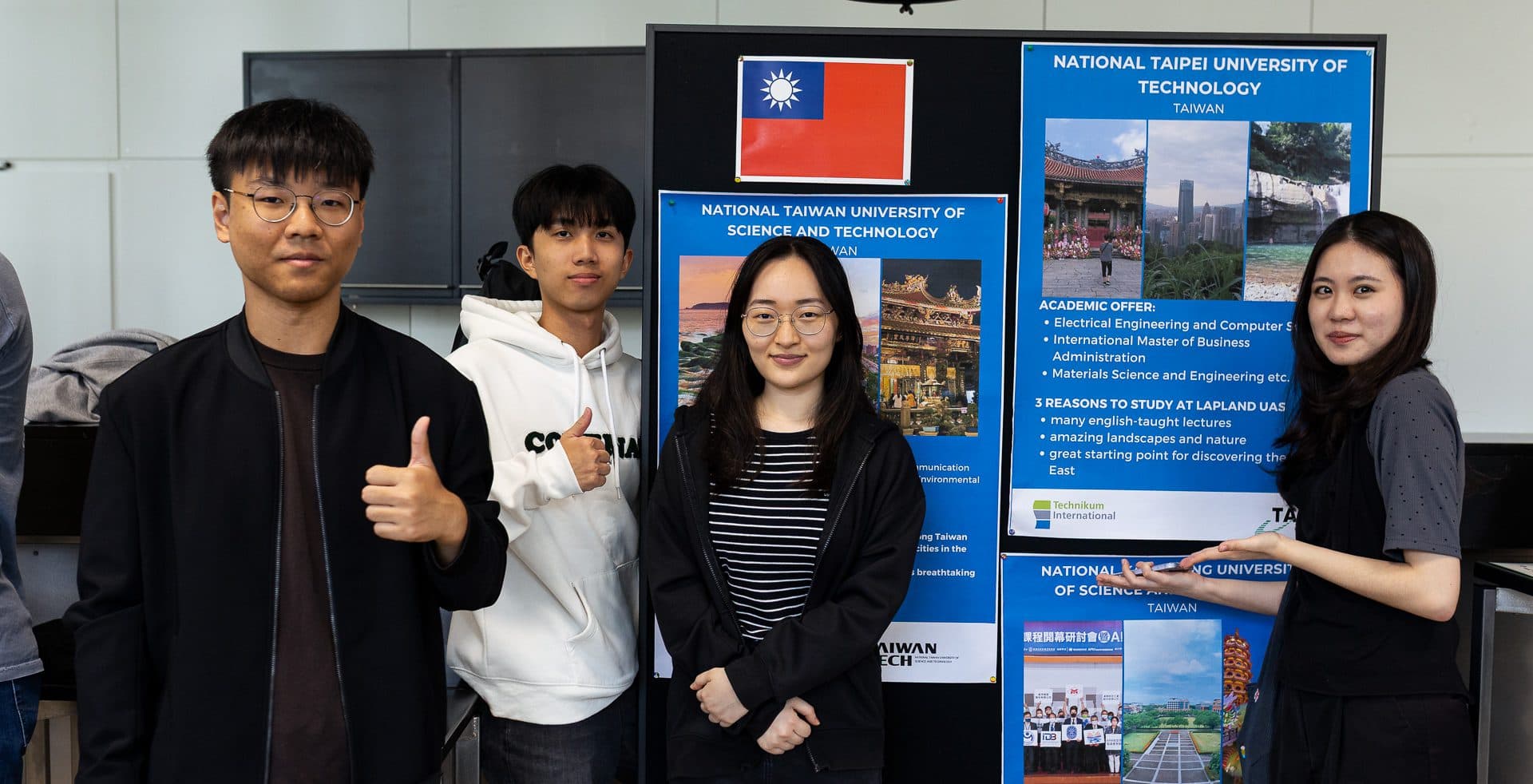 Vier Personen stehen vor Informationsplakaten taiwanesischer Hochschulen, darunter die Nationale Taiwan-Universität für Wissenschaft und Technologie und die Nationale Technische Universität Taipeh. Eine Person zeigt den Daumen nach oben.