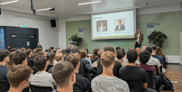Ein Mann steht auf der Bühne und spricht zu den sitzenden Zuhörern. Auf einer Präsentationsfolie hinter ihm sind die Fotos und Namen zweier Personen zu sehen. Die Veranstaltung scheint in einem modernen Hörsaal stattzufinden, an dessen Wänden Plakate und Pflanzen hängen.