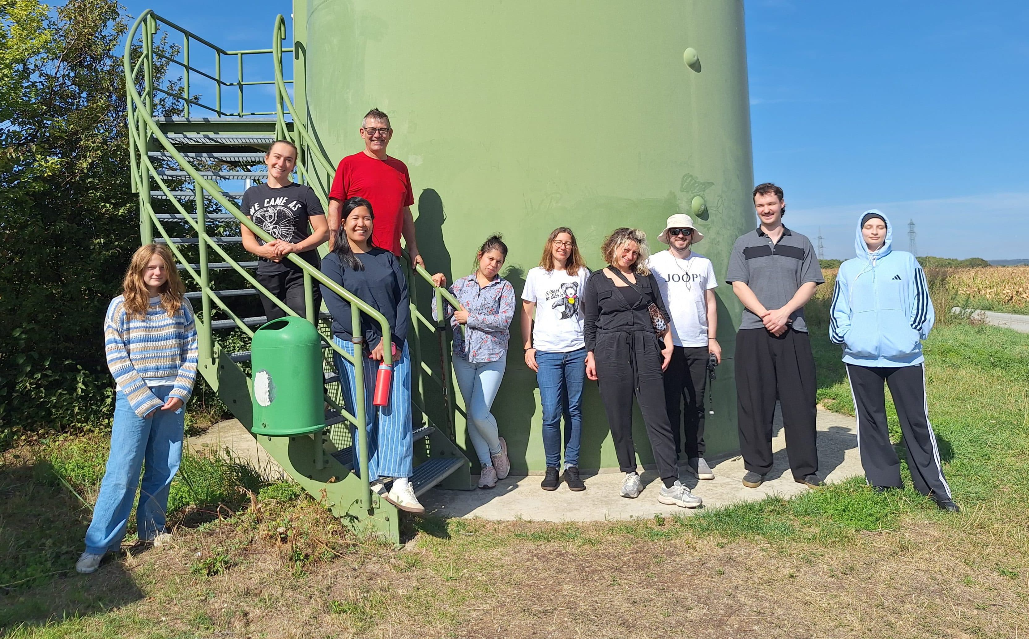 Eine Gruppe von neun Personen, die vor einem großen, hellgrünen Industrieturm mit der Nummer 70112 für ein Foto posieren.