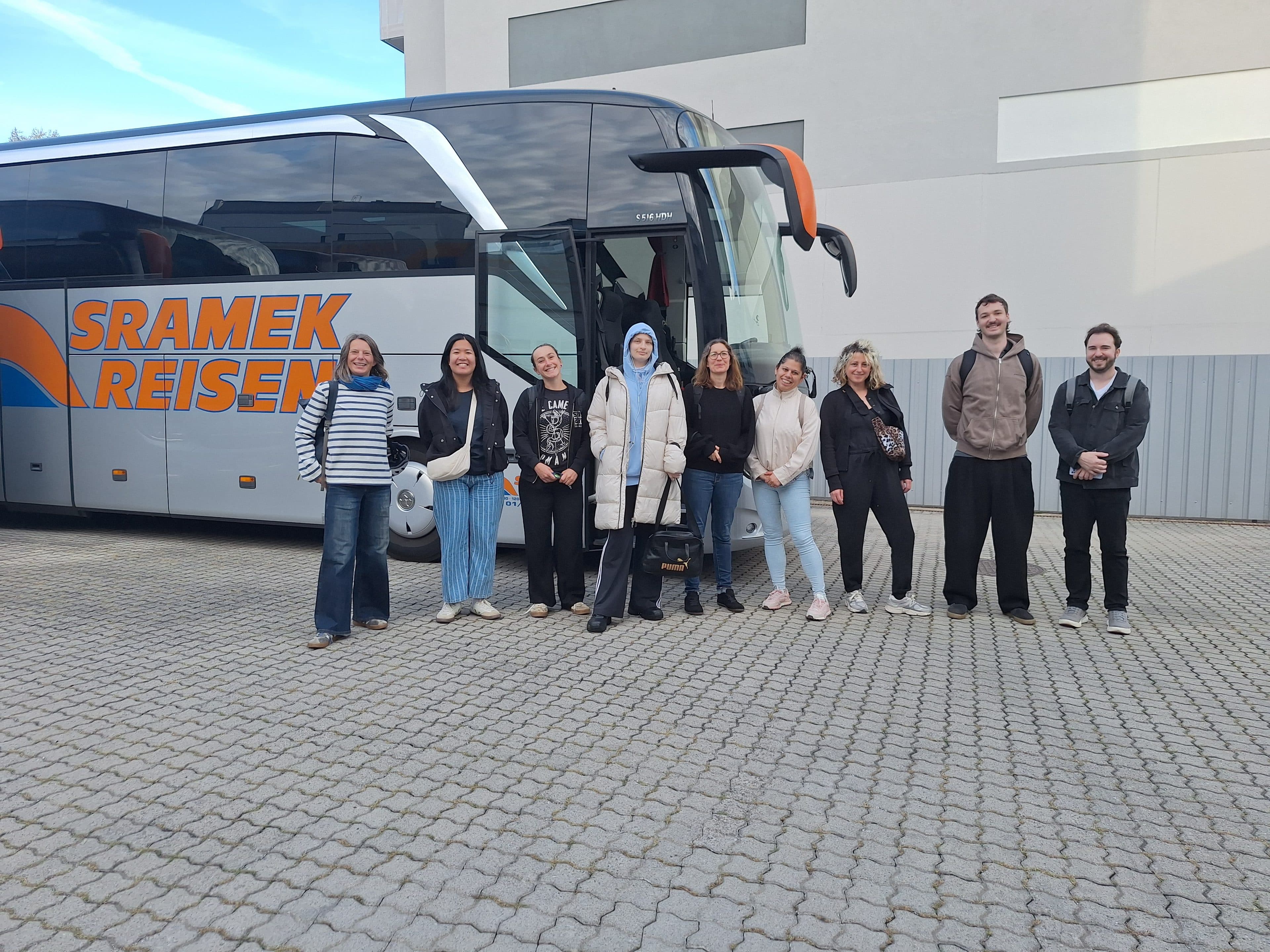 Eine Gruppe von neun Personen steht in einer Reihe auf einem gepflasterten Platz vor einem geparkten Reisebus mit dem Schriftzug Sramek Reisen.
