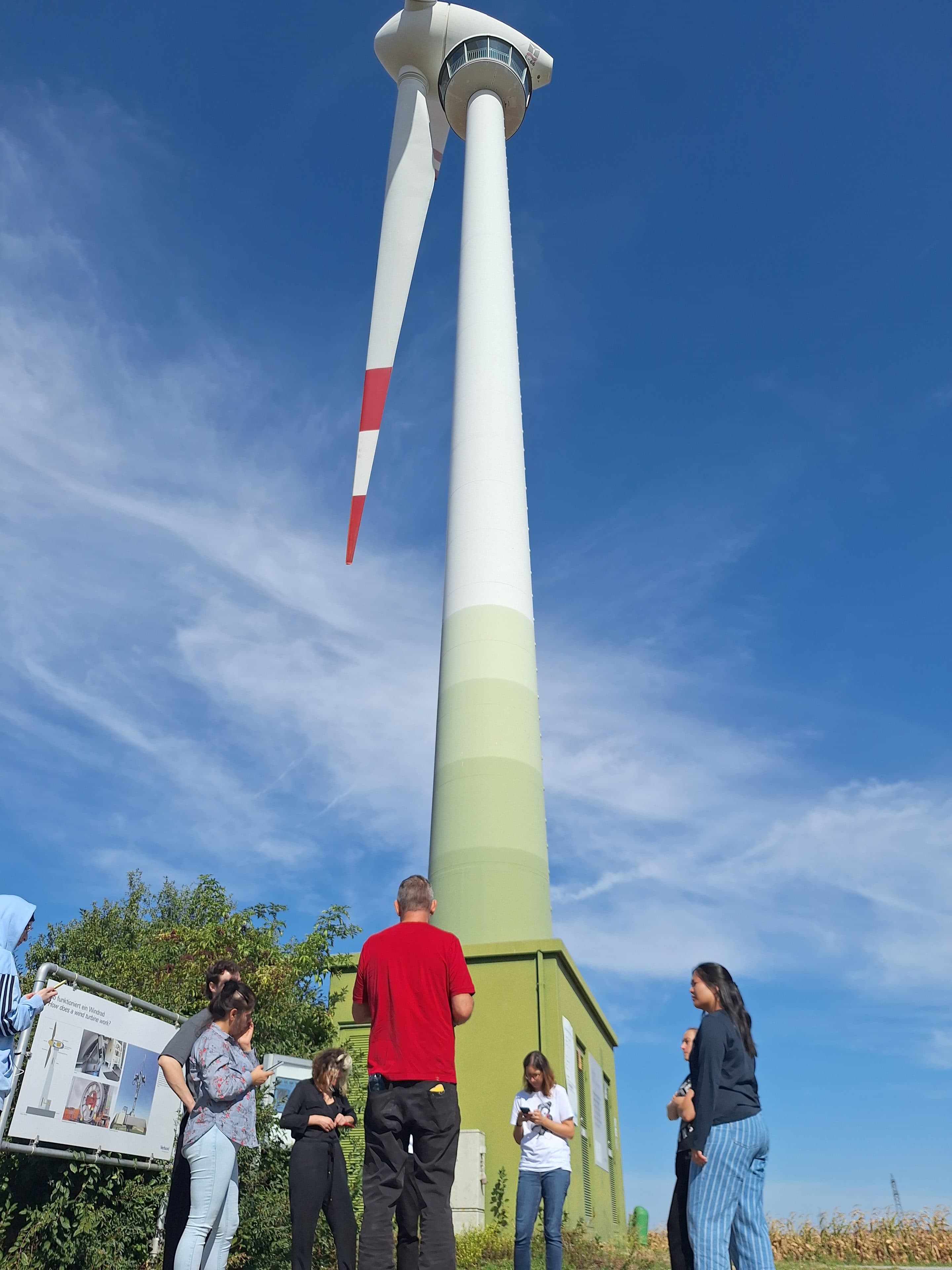 Eine Gruppe von Menschen, die an einem sonnigen Tag am Fuße einer hohen Windkraftanlage steht.