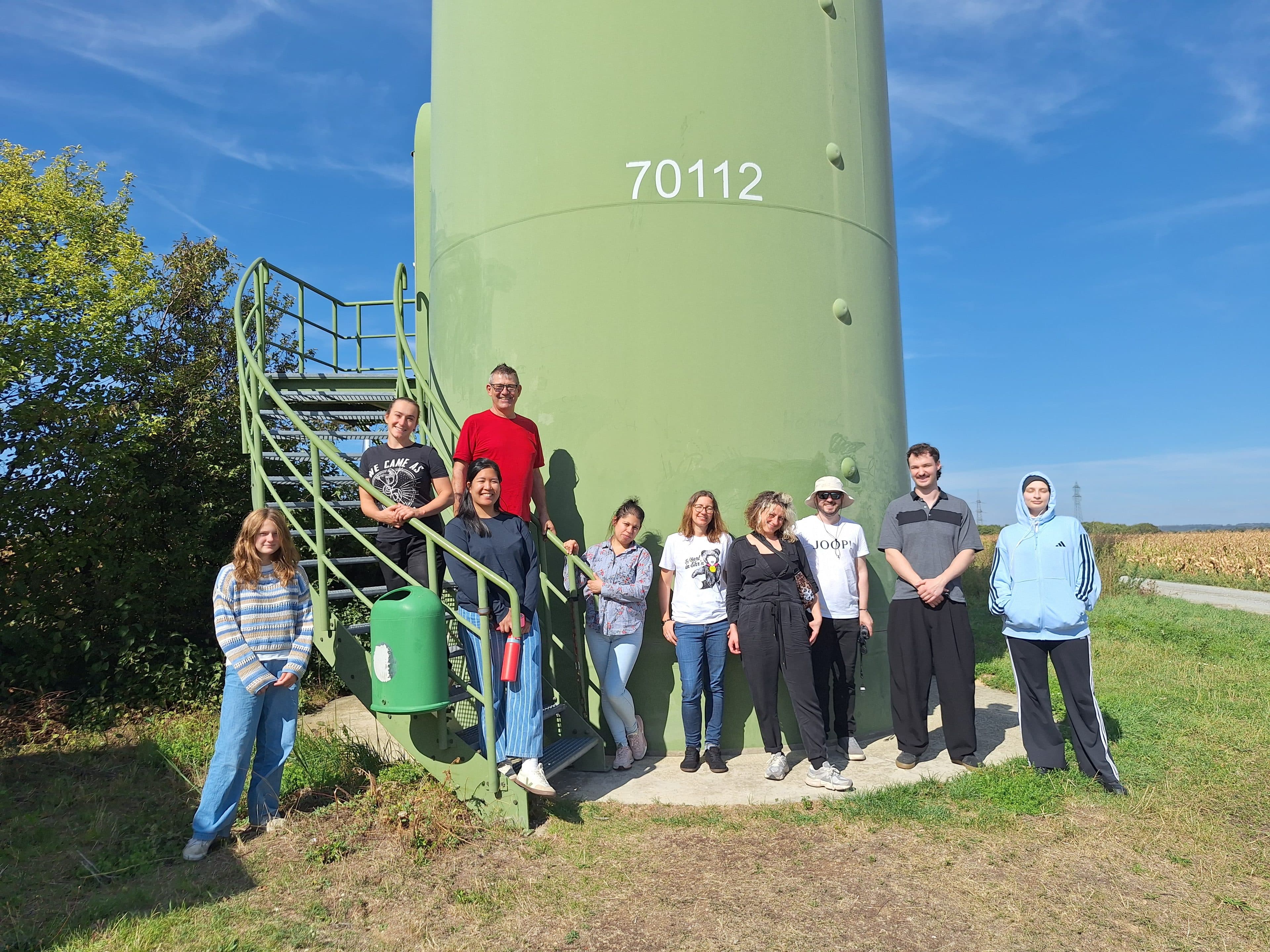Eine Gruppe von neun Personen posiert vor einem großen, hellgrünen Industrieturm, der mit der Nummer 70112 gekennzeichnet ist.