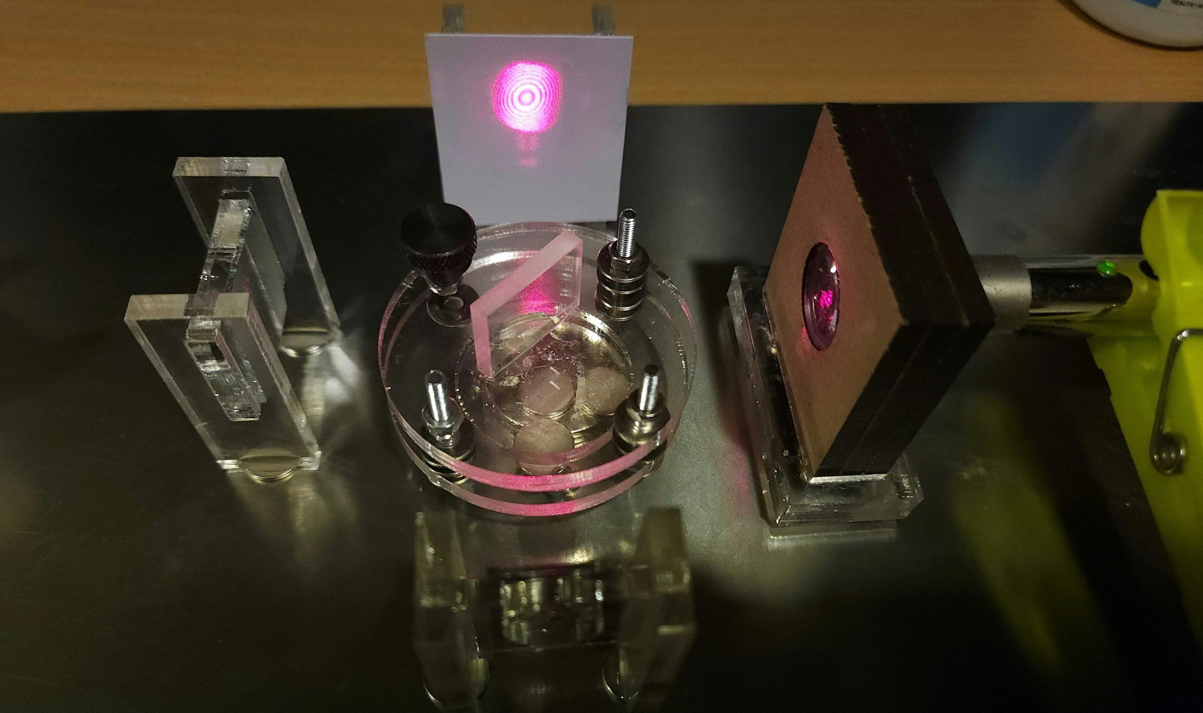 Ein Experimentieraufbau zur Laseroptik auf einer Metalloberfläche, bei dem ein Laserstrahl durch Bauteile hindurchgeht und auf einem Bildschirm ein kreisförmiges Interferenzmuster erzeugt.