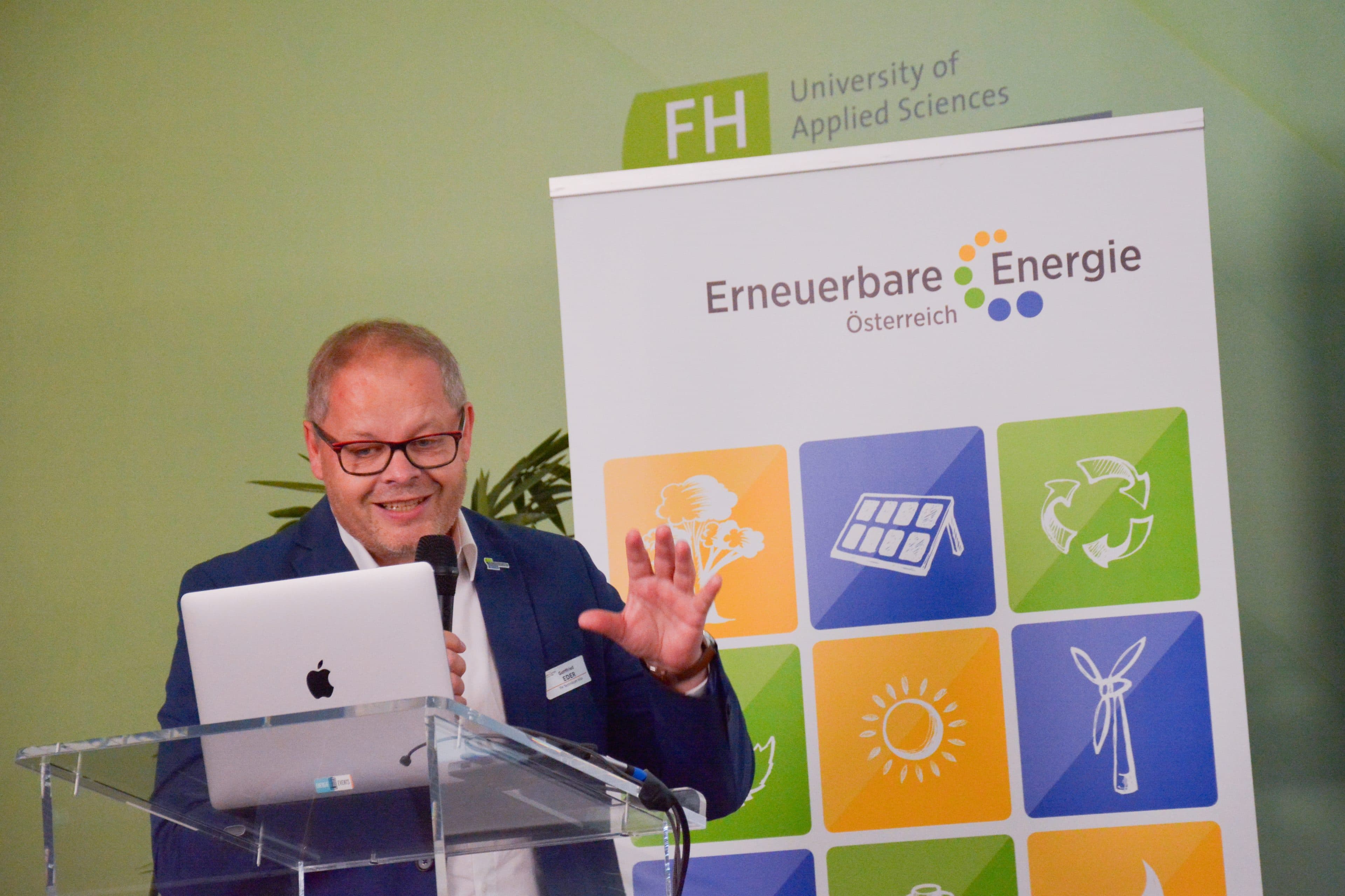 Ein Mann in einem Anzug spricht an einem Rednerpult vor einem Banner mit der Aufschrift Erneuerbare Energie, auf dem Symbole für erneuerbare Energiequellen abgebildet sind.