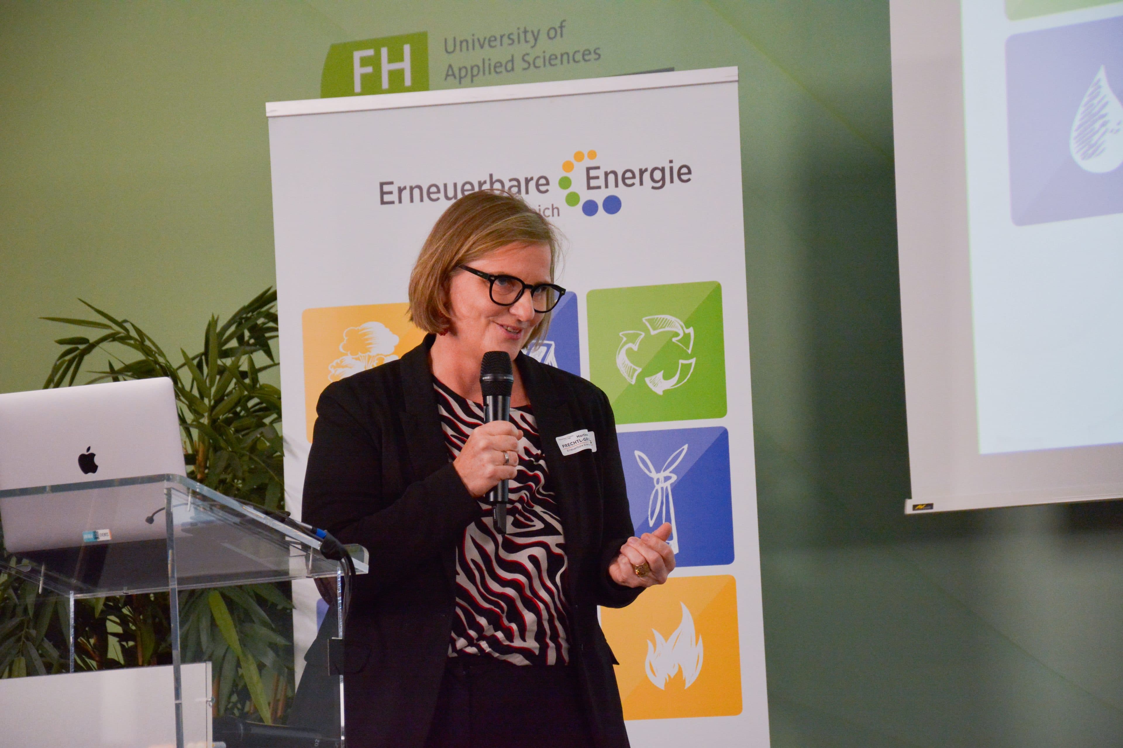 Eine Frau mit Brille spricht an einem Rednerpult in ein Mikrofon. Hinter ihr ist ein Banner der FH University of Applied Sciences zu sehen, auf dem der Studiengang Erneuerbare Energie mit Symbolen für Wind, Recycling und Feuer abgebildet ist.