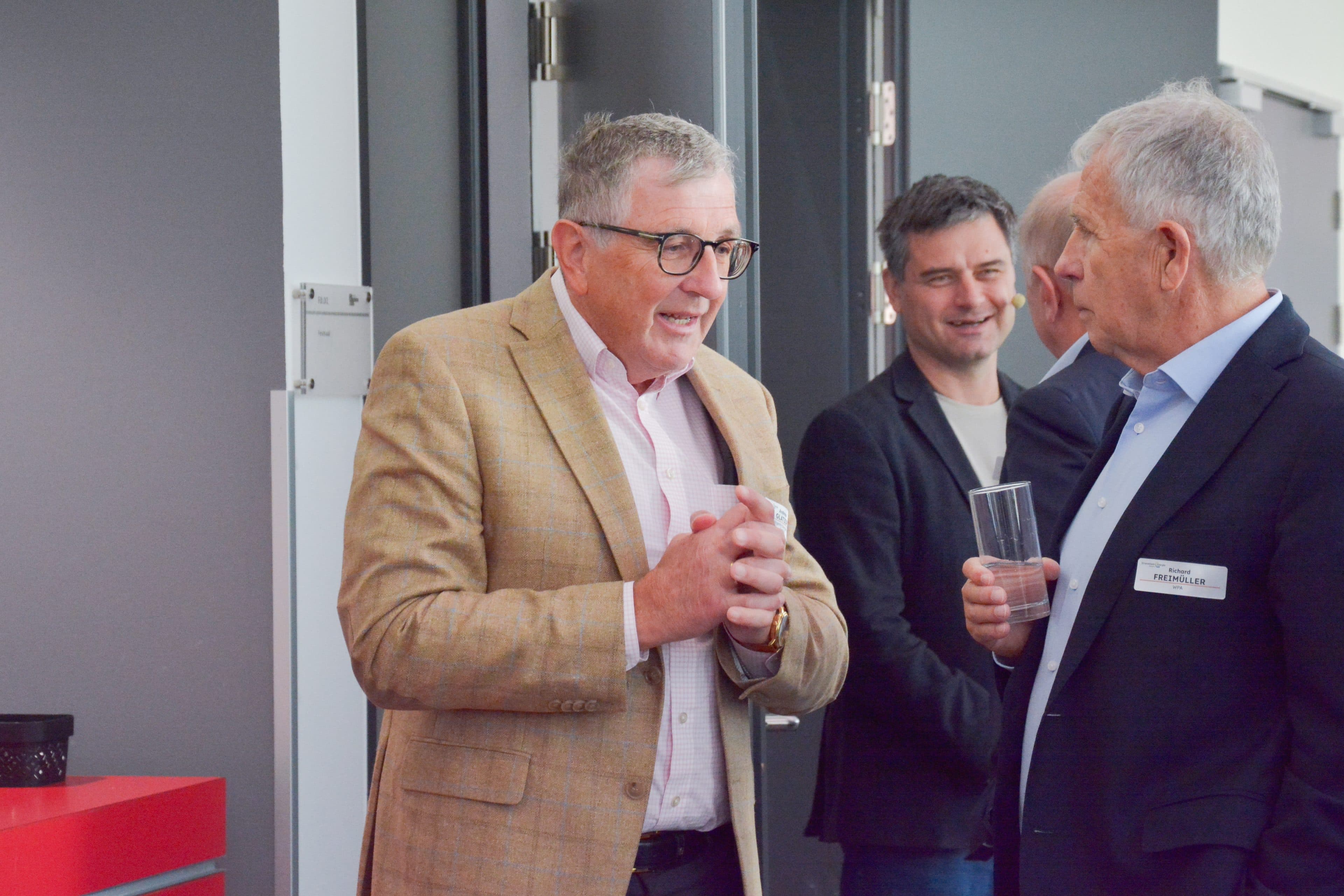 Drei Männer unterhalten sich bei einer Veranstaltung. Ein Mann in einem beigen Blazer und mit Brille spricht mit Richard Freimüller, der ein Getränk in der Hand hält. Ein dritter Mann lächelt im Hintergrund.