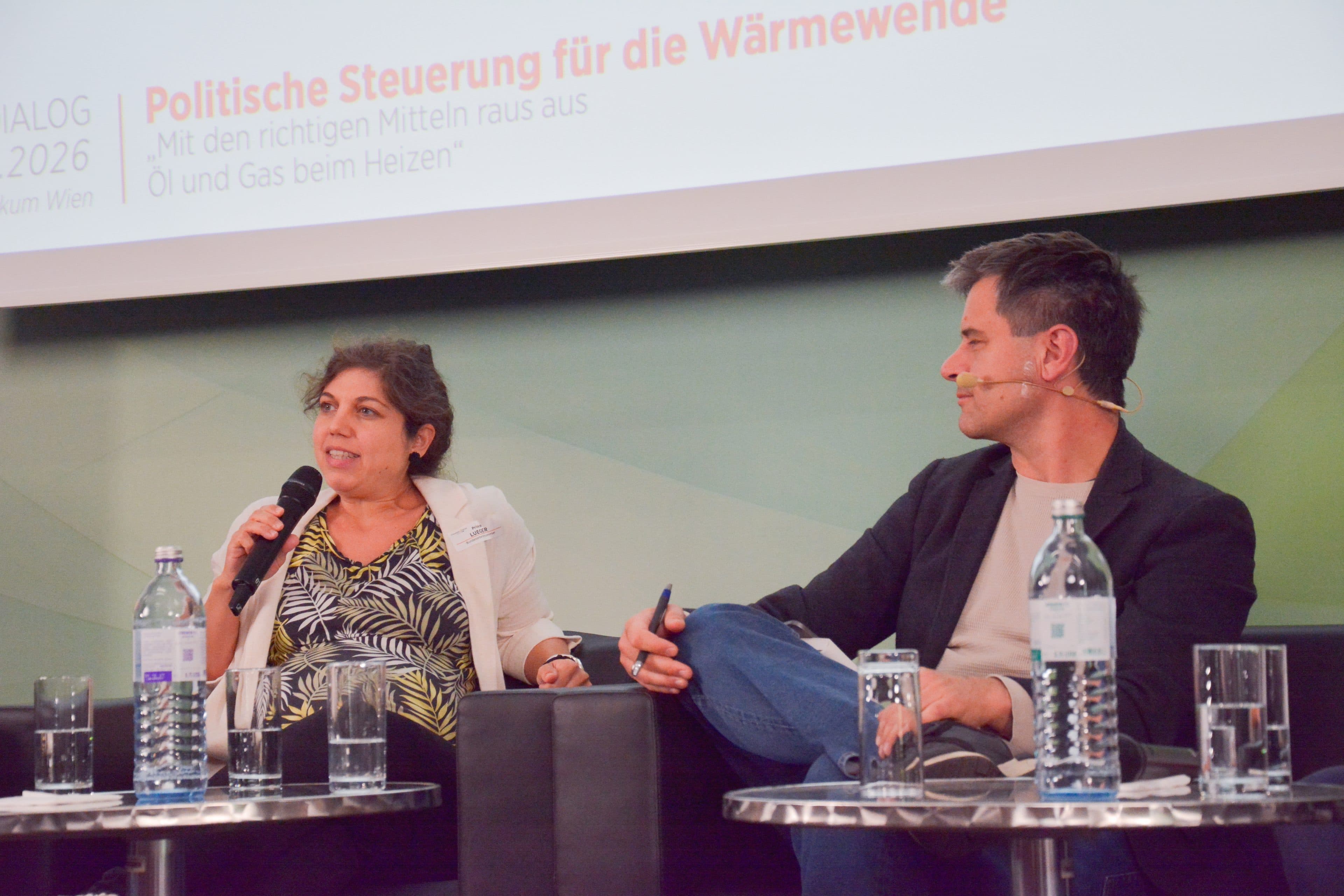 Zwei Personen sitzen mit Mikrofonen auf der Bühne einer Podiumsdiskussion. Die Frau auf der linken Seite spricht und gestikuliert dabei mit der rechten Hand. Der Mann auf der rechten Seite hört zu und lächelt. Auf dem Tisch vor ihnen stehen Wasserflaschen und Unterlagen.