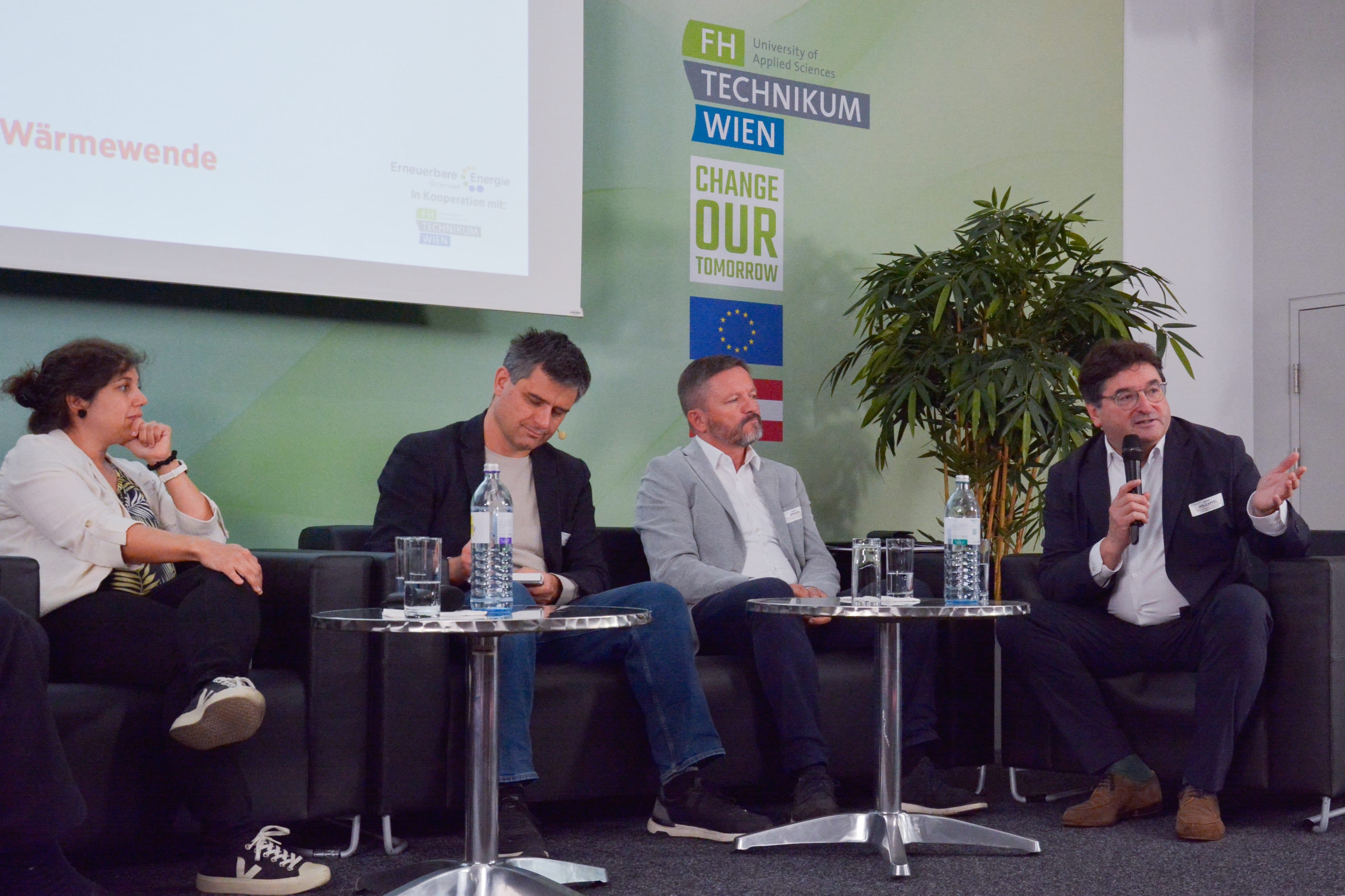 Vier Personen sitzen während einer Podiumsdiskussion auf einer Bühne, wobei eine Person in ein Mikrofon spricht.