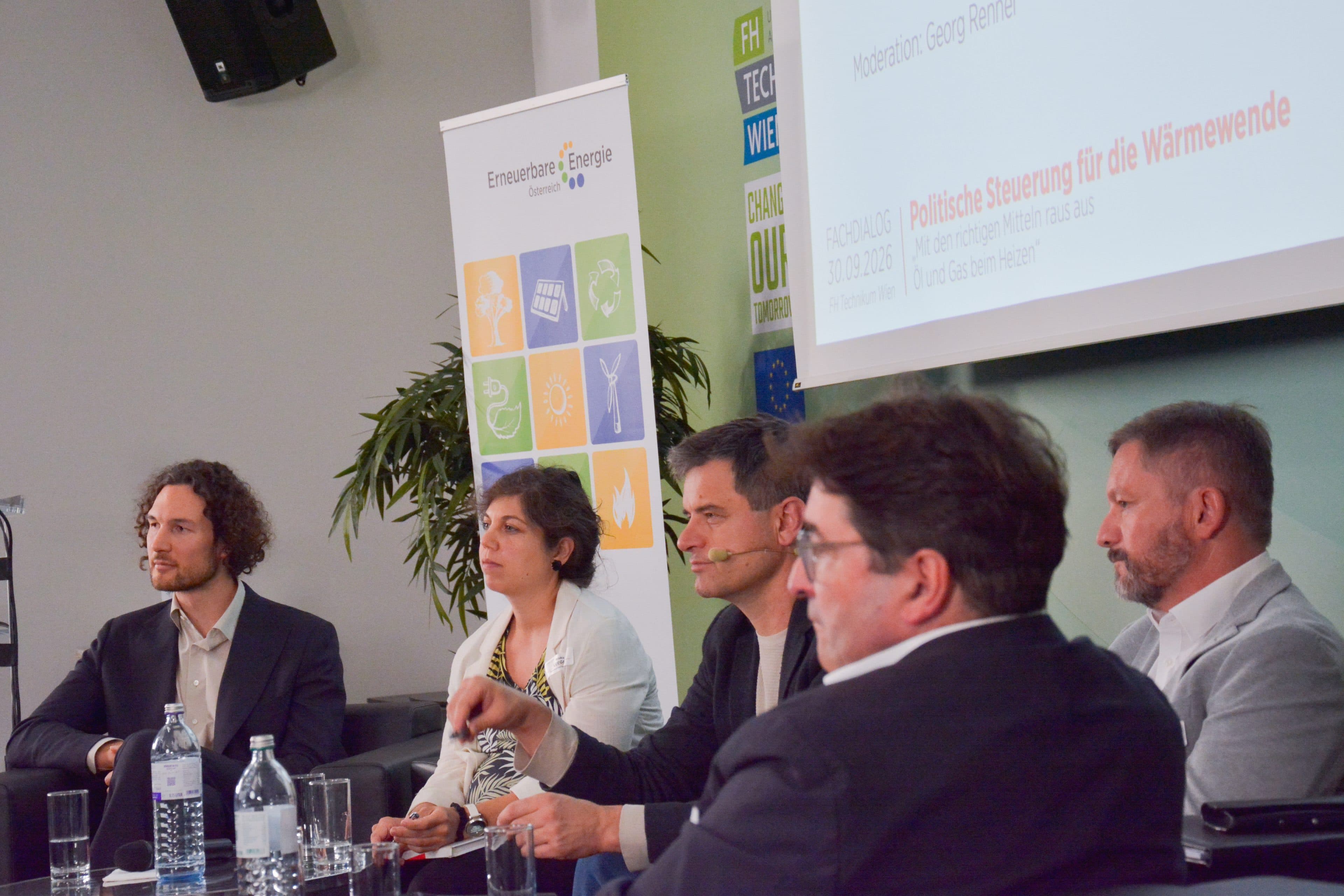 Fünf Personen sitzen an einem Podiumsdiskussions-Tisch mit Mikrofonen und Wasserflaschen, im Hintergrund sind eine Präsentationsleinwand und ein buntes Banner zum Thema Energie zu sehen. Die Kulisse lässt auf eine Konferenz oder ein Seminar zur Energiewende schließen.