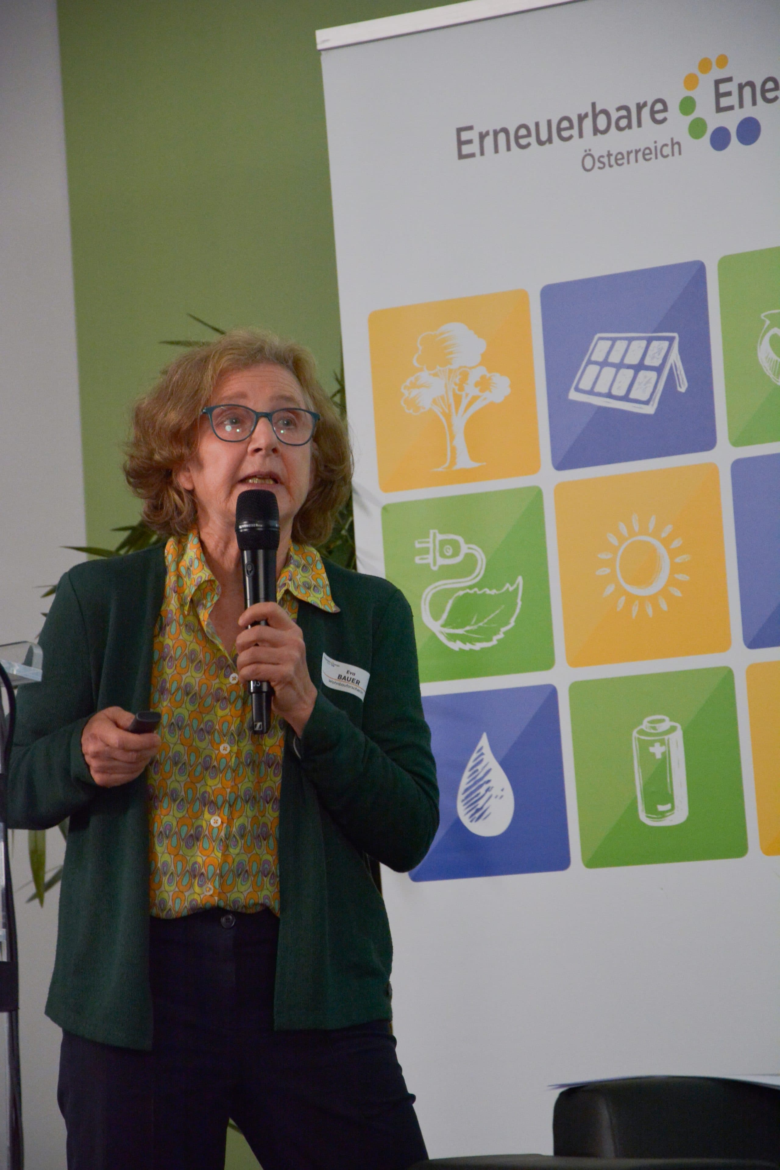 Eine Frau in einer gemusterten Bluse und einer grünen Strickjacke spricht vor einem Werbebanner von Erneuerbare Energie Österreich in ein Mikrofon.
