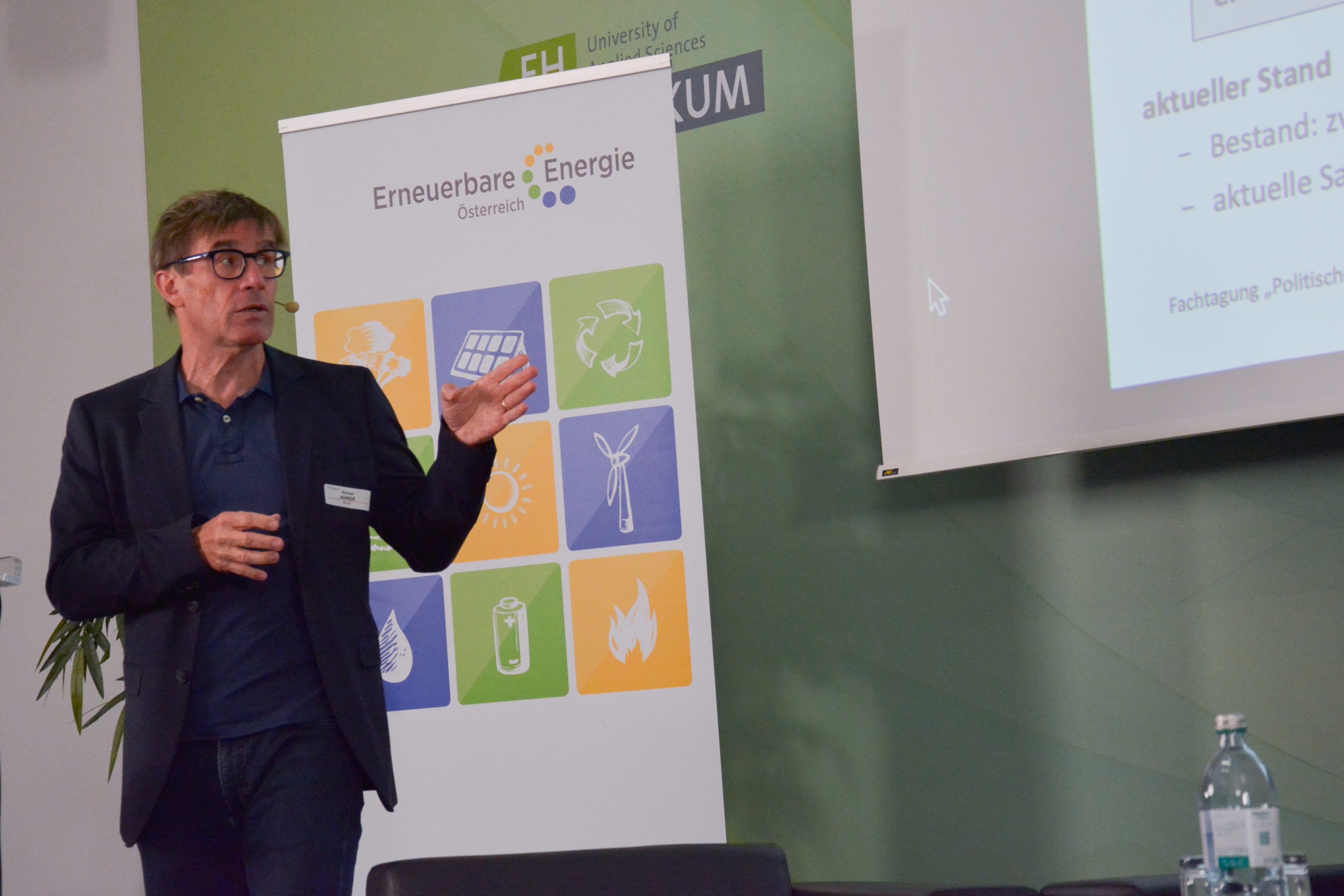 Ein Mann in einem Blazer gestikuliert, während er vor einem Banner mit der Aufschrift Erneuerbare Energie und einer projizierten Präsentation spricht.