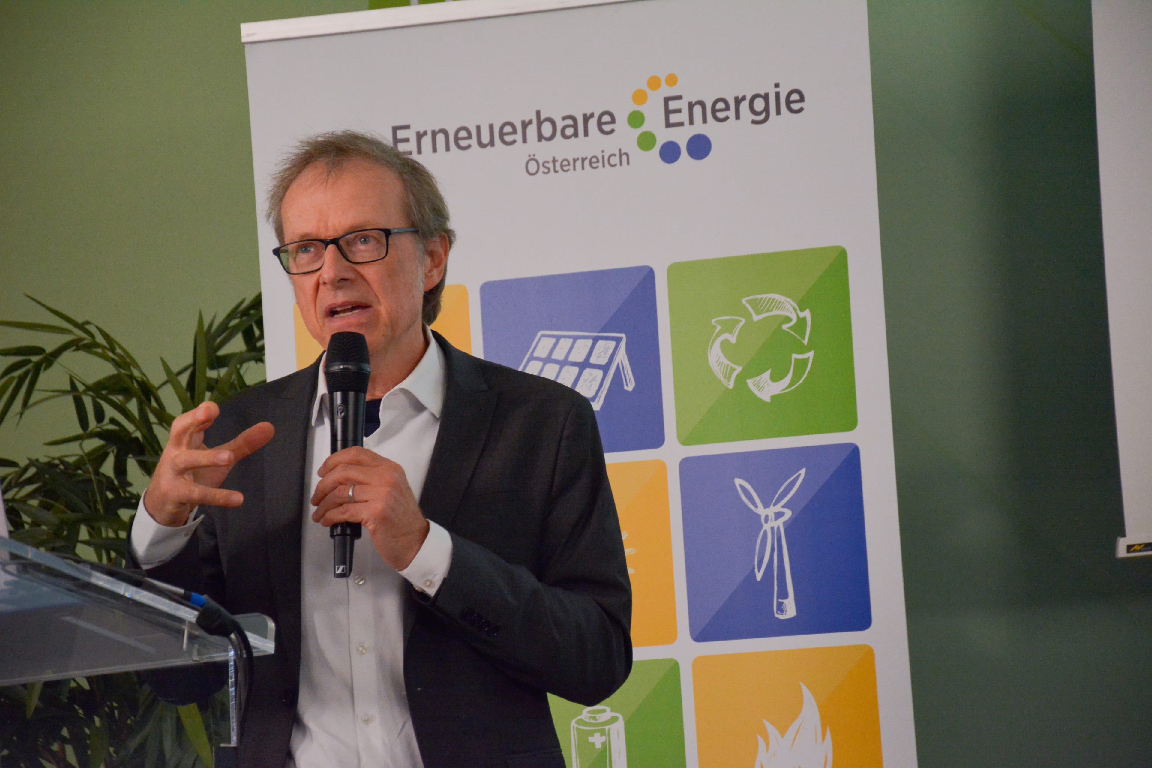 Ein Mann im Anzug und mit Brille spricht vor einem Banner von Erneuerbare Energie Österreich, auf dem Umweltsymbole abgebildet sind, in ein Mikrofon.