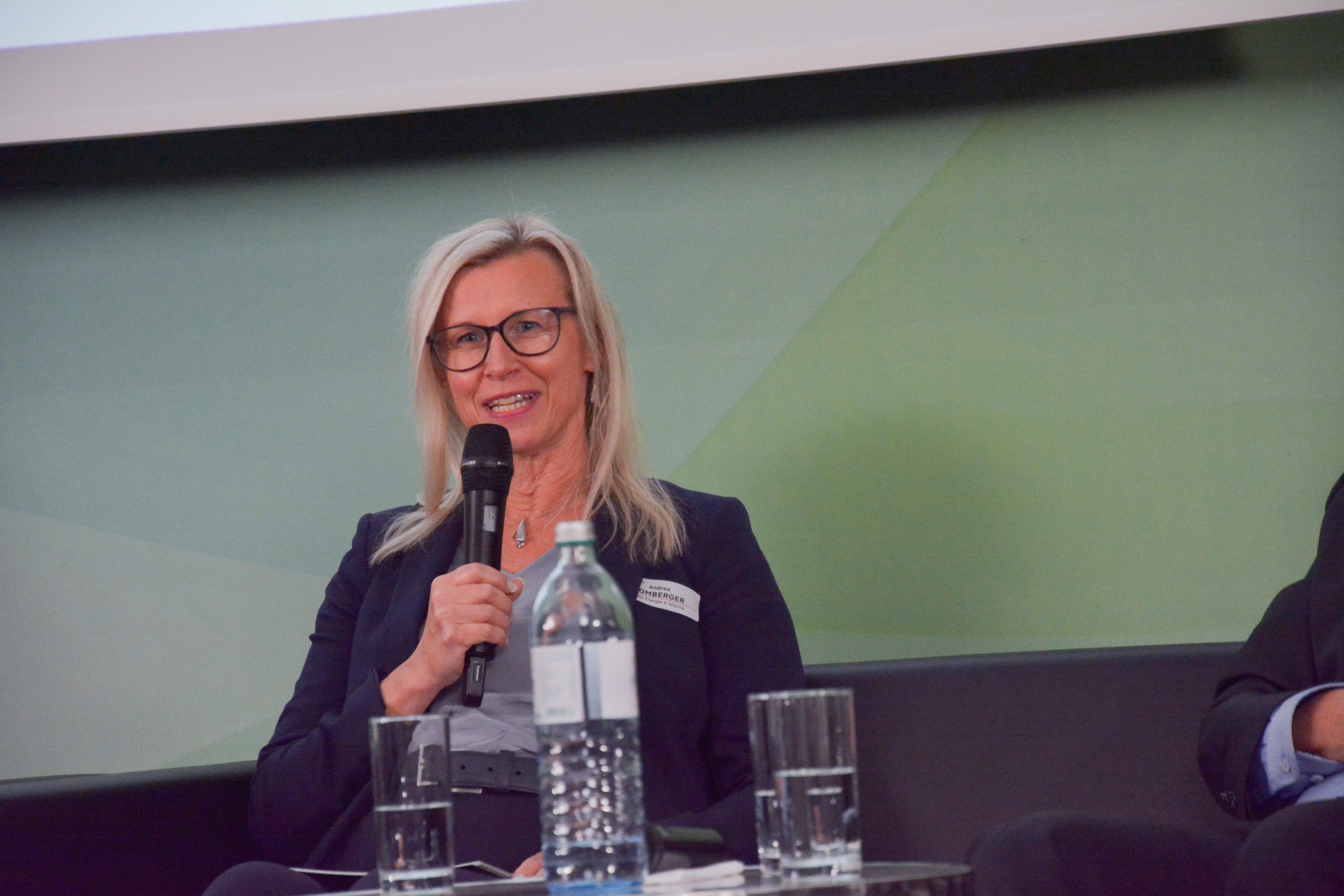 Eine Frau mit blonden Haaren und Brille spricht bei einer Podiumsdiskussion lächelnd in ein Mikrofon. Sie trägt einen dunklen Blazer mit einem Namensschild. Auf dem Tisch vor ihr stehen Wasserflaschen und Gläser.